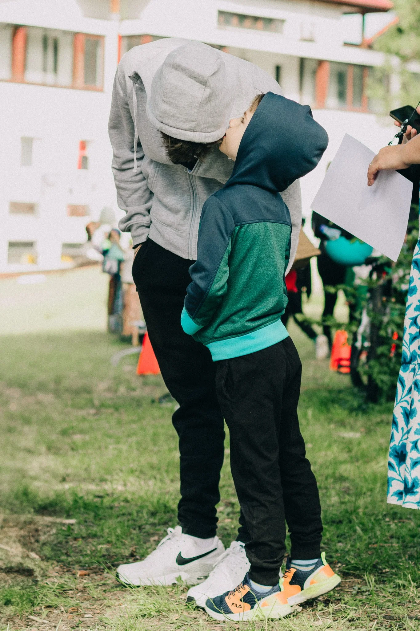 An adult and a child are standing outdoors, with the adult leaning down and touching the child's face. The adult is wearing a gray hoodie and black pants, and the child is in a green and blue jacket with black pants and sneakers. A person on the righ