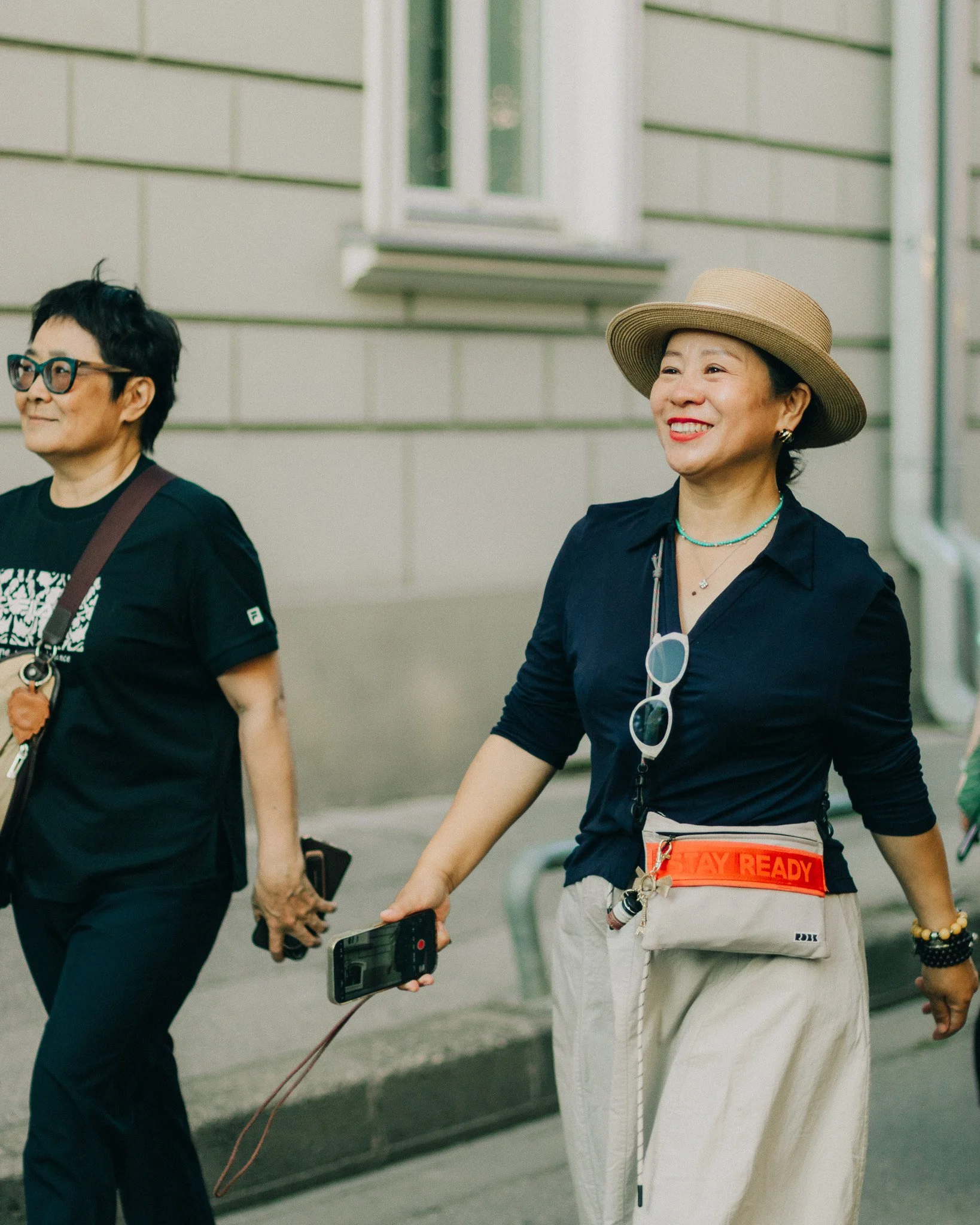 Two women walking outdoors, one smiling with a wide-brimmed hat and sunglasses hanging from her shirt, the other wearing glasses and a black t-shirt, holding a phone.