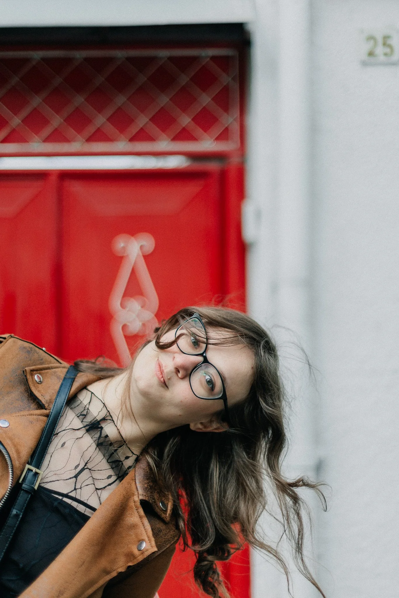 A young woman with long, curly hair and glasses tilting her head in front of a red wall or door.