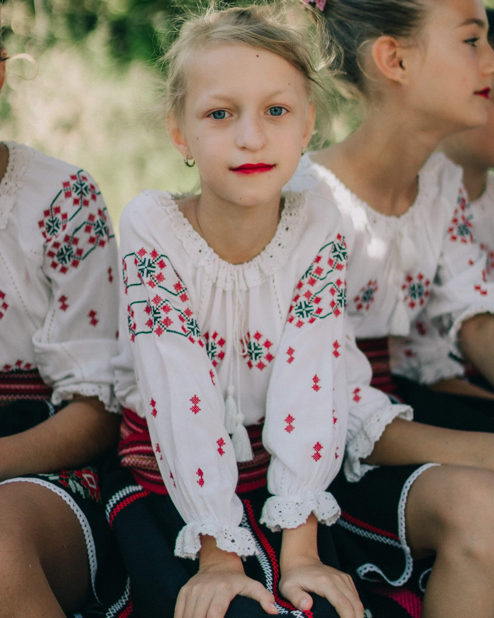 A girl wearing traditional embroidered white blouse and black skirt, sitting outdoors with other girls dressed similarly.