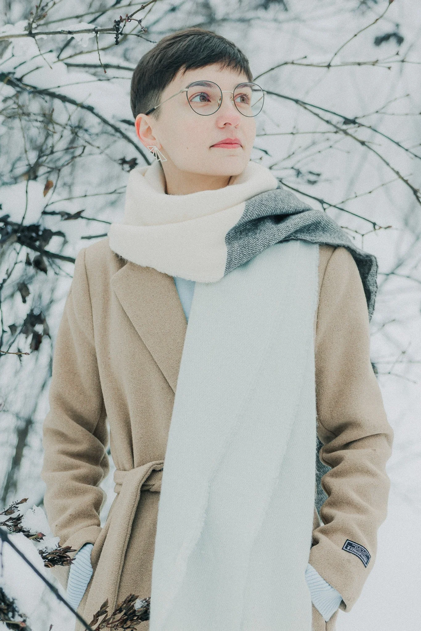 A young person with short dark hair, glasses, and earrings stands outdoors in a snowy landscape with bare tree branches in the background. They are wearing a tan coat, a white scarf, and a white and gray scarf.