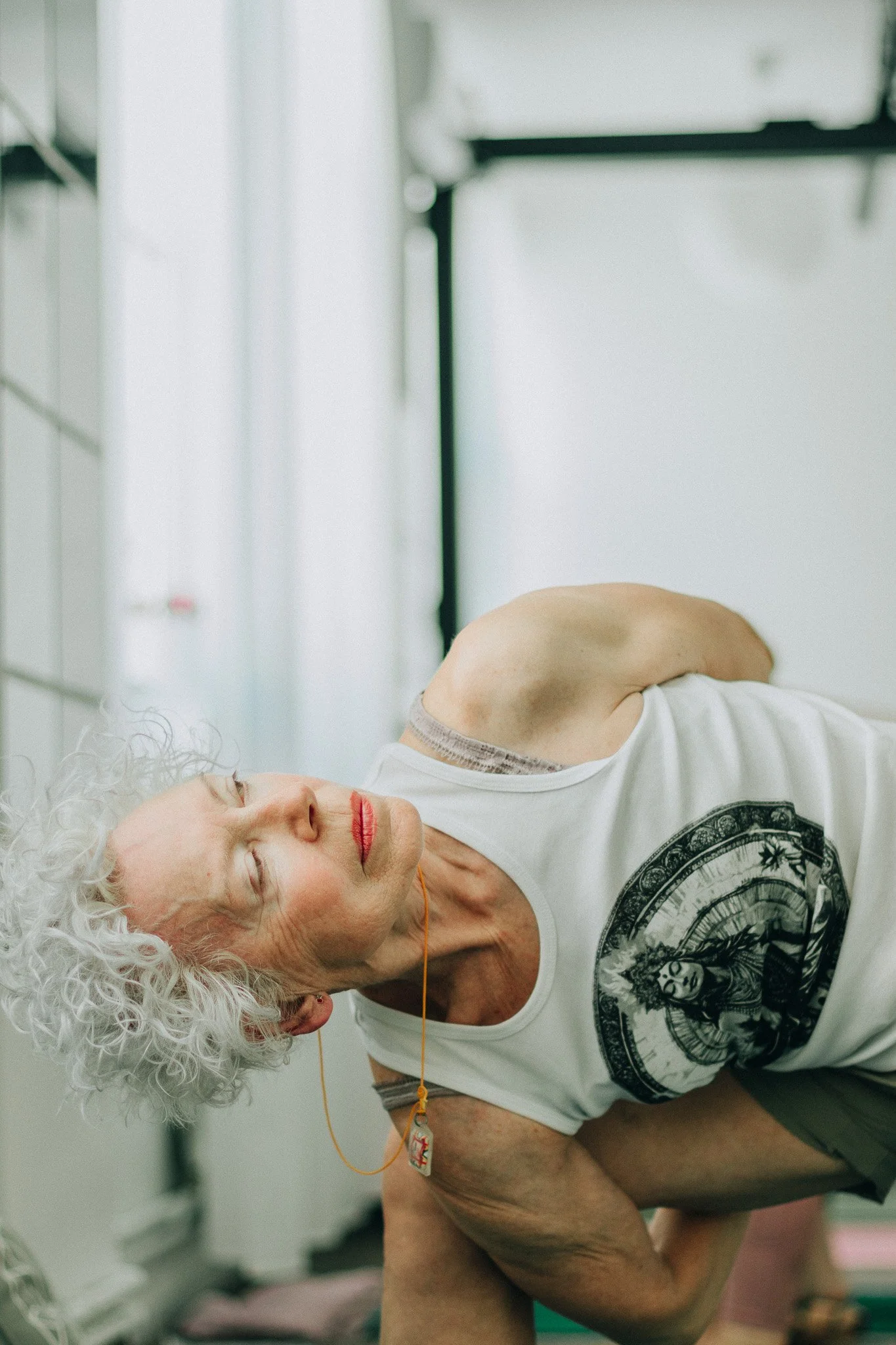 An elderly woman with curly white hair practicing yoga or stretching, with eyes closed and a calm expression, in a bright indoor space.