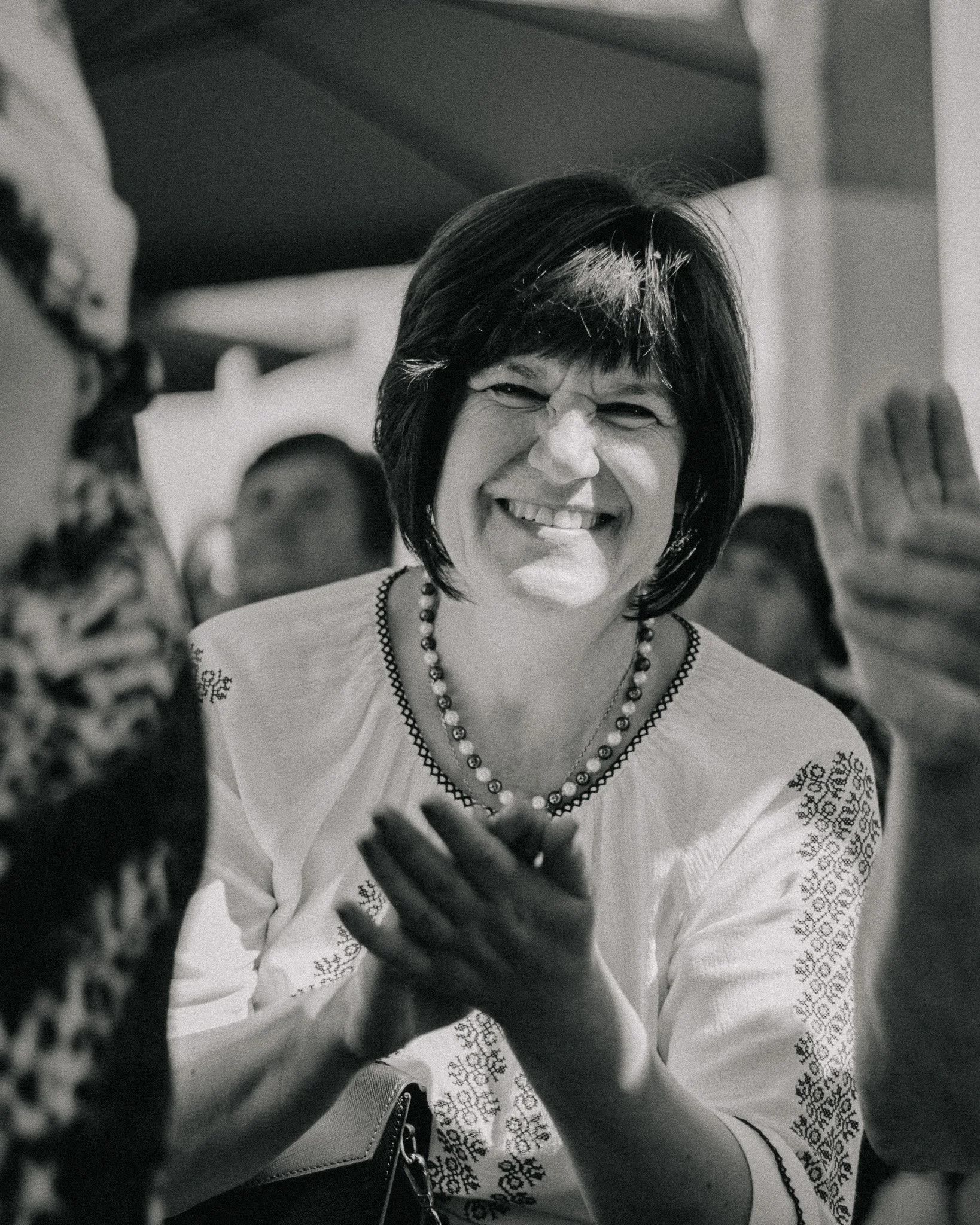 A woman with short dark hair smiling brightly, wearing a white embroidered blouse and a beaded necklace, in a social setting.