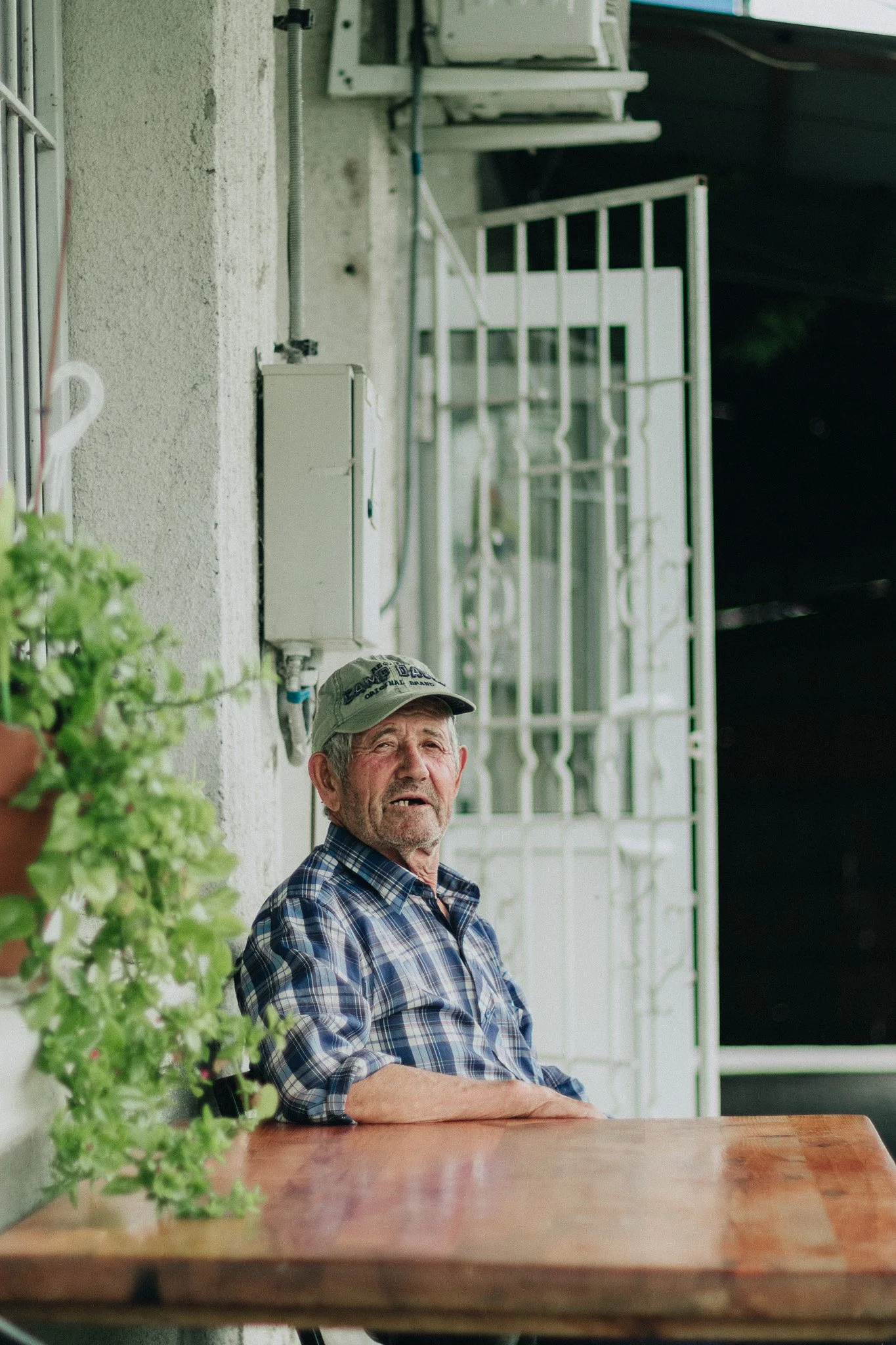 An elderly man sitting at a wooden table on a porch, wearing a plaid shirt and cap, with a potted plant and a white metal security gate in the background.