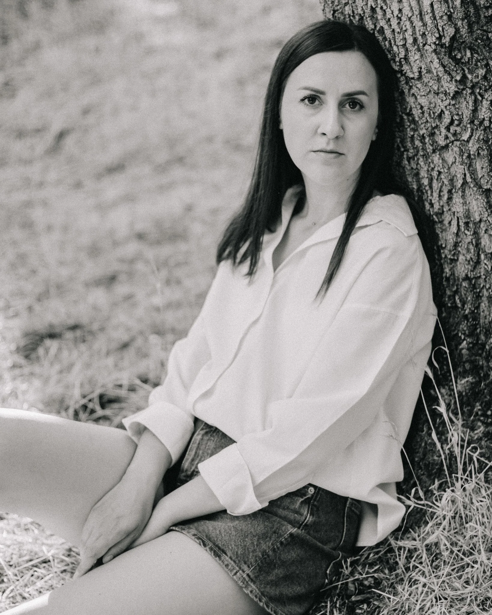 A woman with long dark hair is sitting on the grass, leaning against a tree trunk, and has a serious expression.