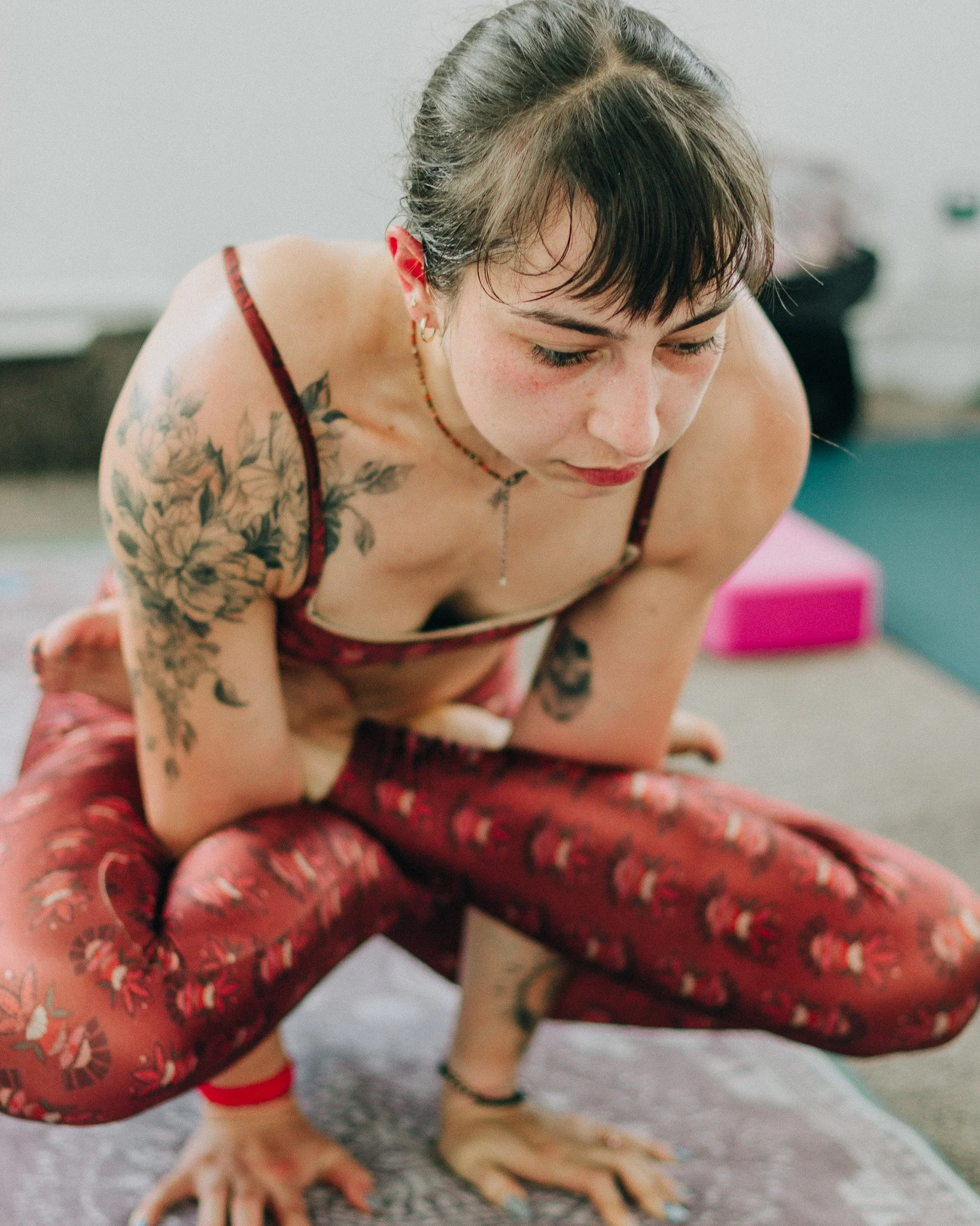 A young woman practicing yoga or stretching on a mat, wearing red patterned leggings and a matching top, with tattoos on her arms and chest, in a bright indoor space.
