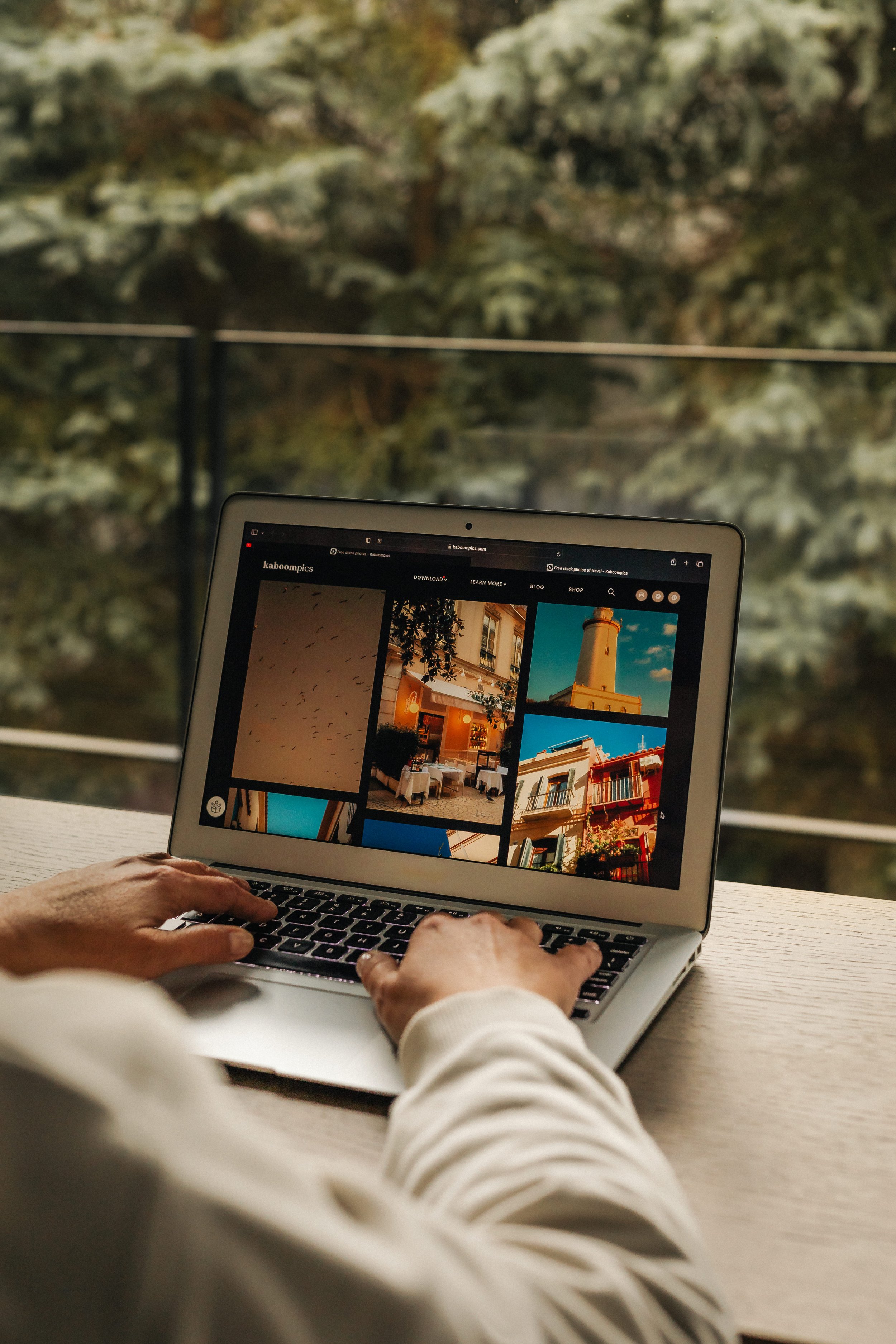 Person using a laptop at a wooden table, browsing images of buildings and scenery, with a blurred outdoor background of trees and foliage.