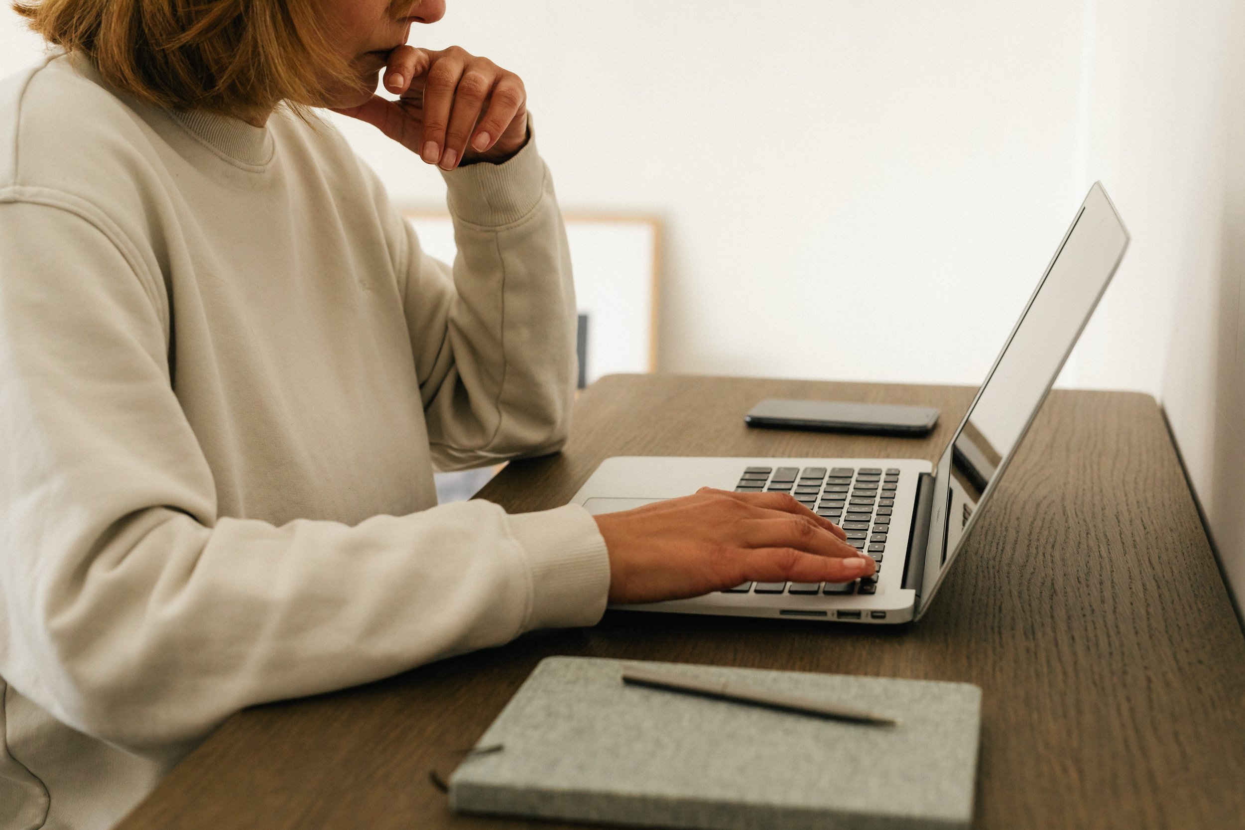 Person in beige sweatshirt typing on a silver laptop at a wooden desk, with a closed notebook and pen in the foreground.
