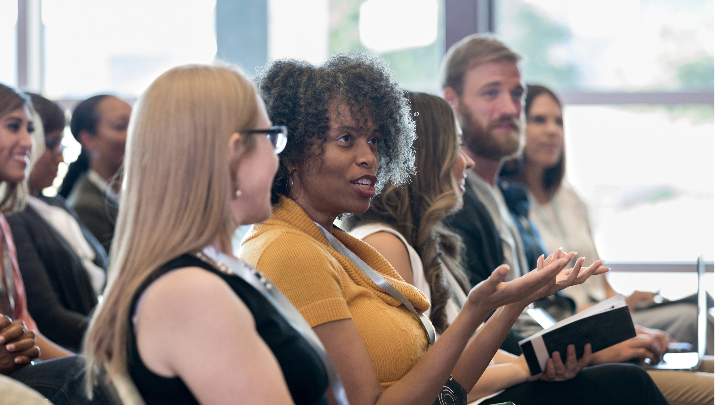 Group of people in a conference or seminar, with a woman in a yellow top speaking while holding a notebook.