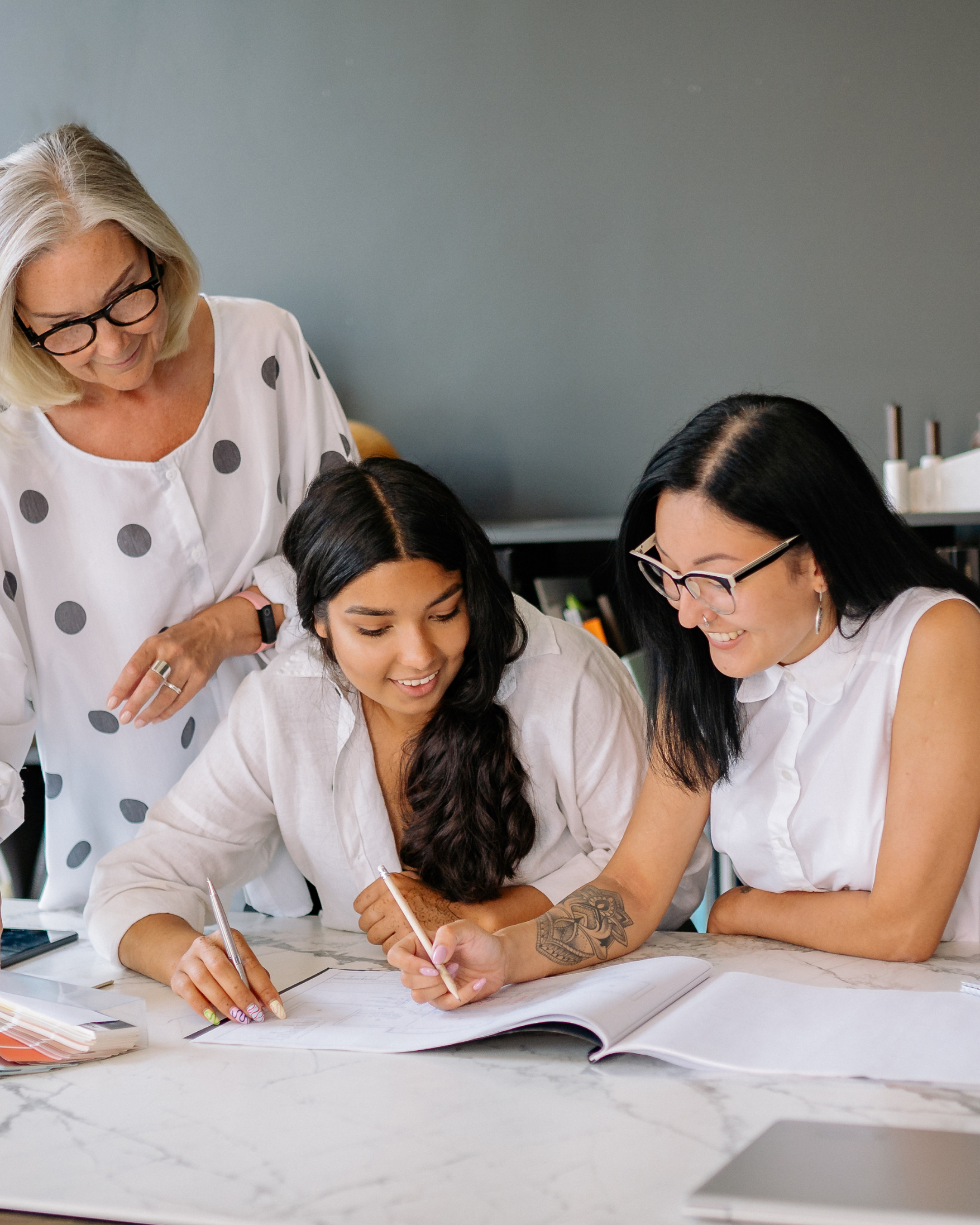 Three women working together at a table, looking at a notebook and smiling