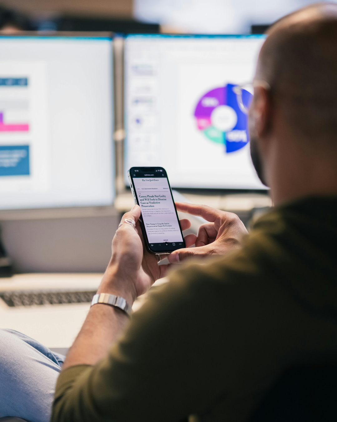 Person with glasses and a watch viewing a smartphone, with dual computer monitors displaying colorful charts and graphs in the background.
