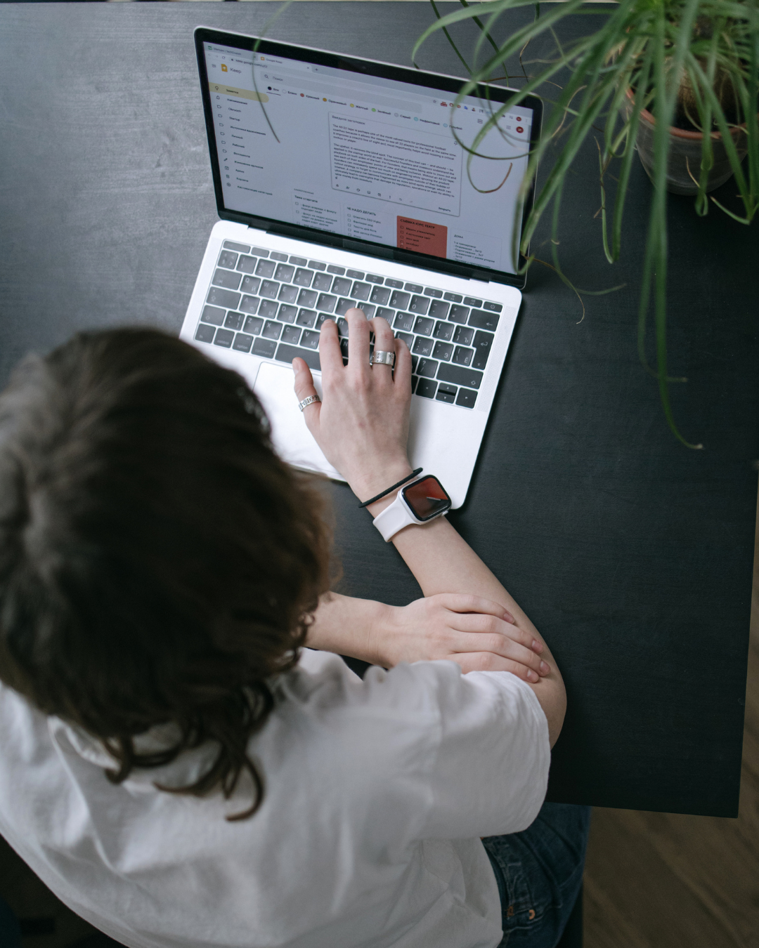 A person with long brown hair working on a silver laptop at a black desk with a potted plant nearby, wearing a white shirt, a smartwatch, and rings.