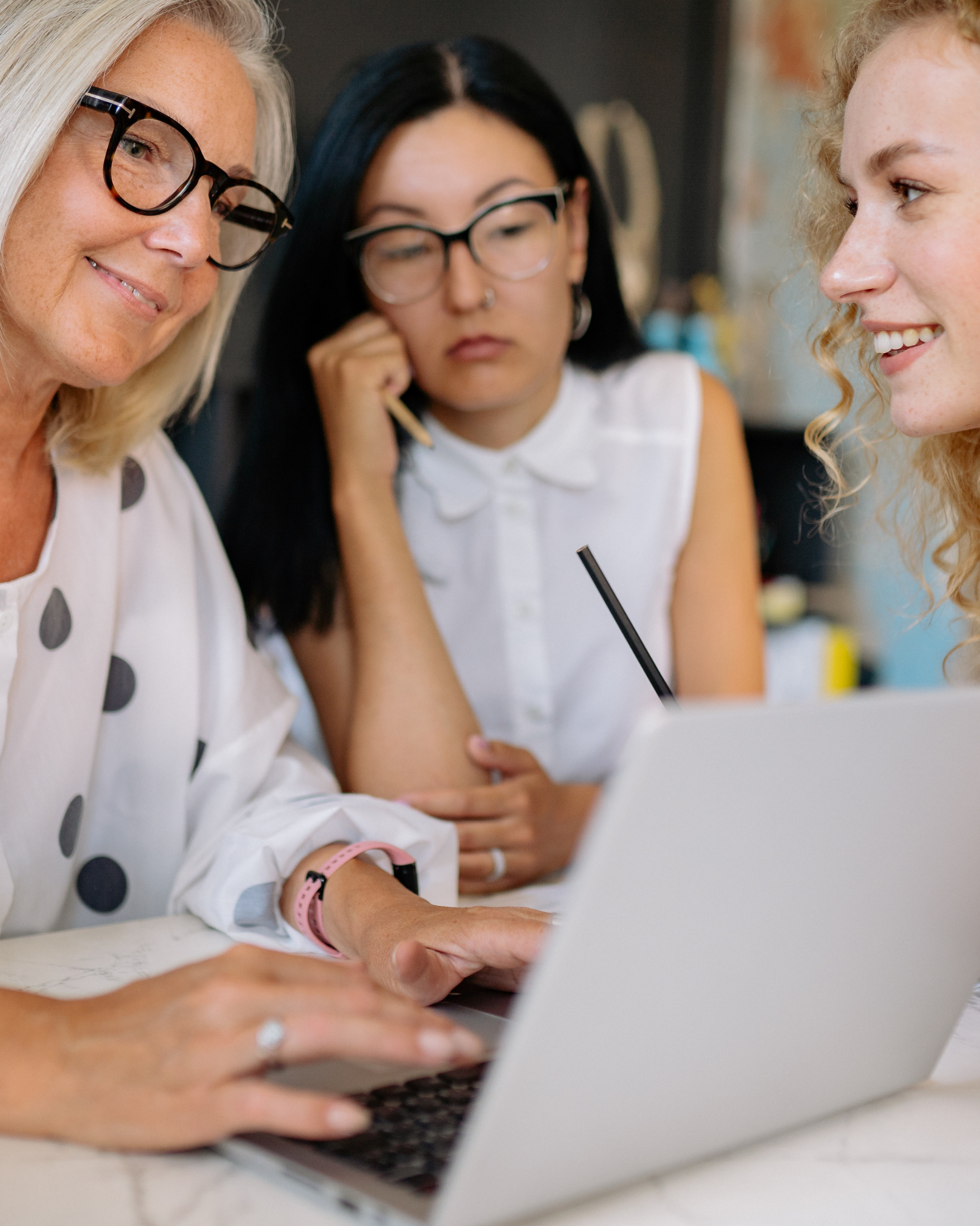 Three women sitting at a table, engaged with a laptop. One older woman with blonde hair and glasses wears a white top with black polka dots; the young woman in the middle has black hair, glasses, and a white sleeveless blouse; the young woman on the right has curly blonde hair and is smiling.