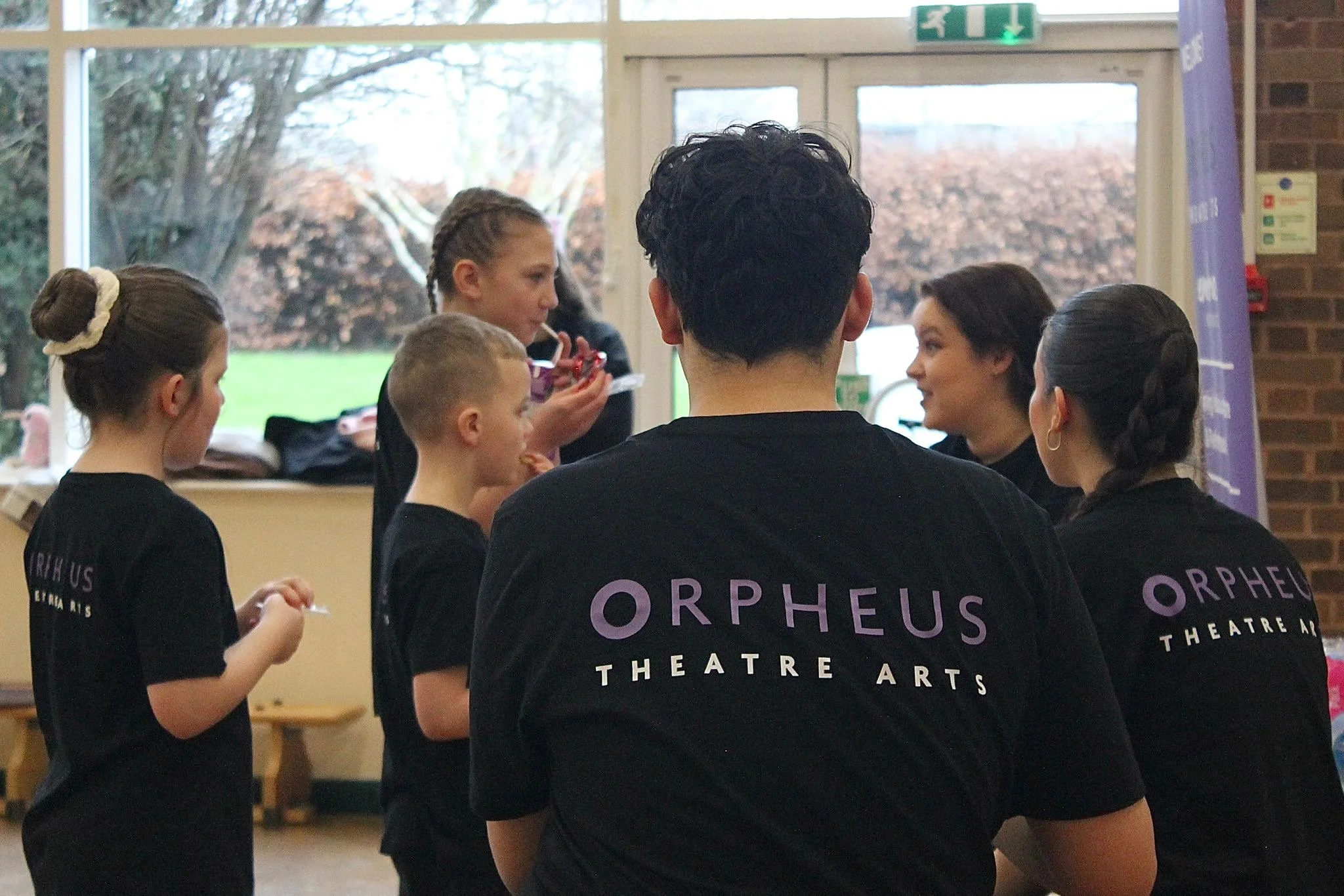 Children and a young adult indoors, all wearing black t-shirts with purple and white text, standing and talking to each other, with large windows showing trees outside.