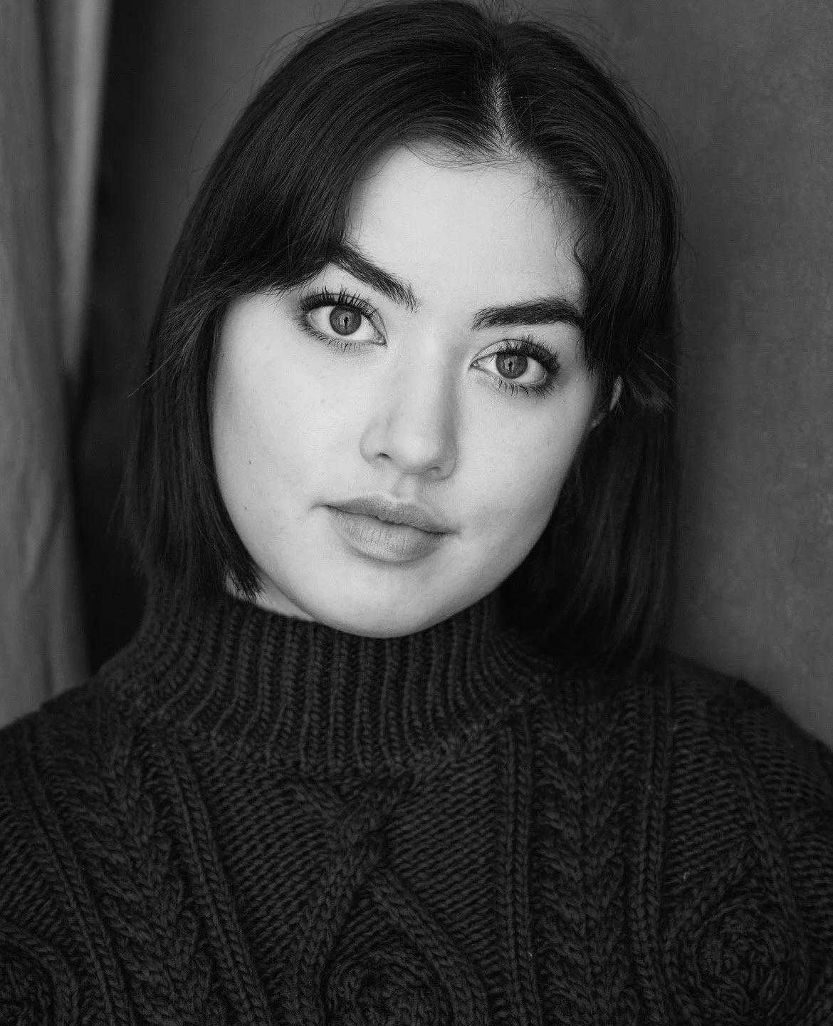 Black and white portrait of a woman with shoulder-length dark hair, wearing a dark knitted sweater, looking directly at the camera with a neutral expression.