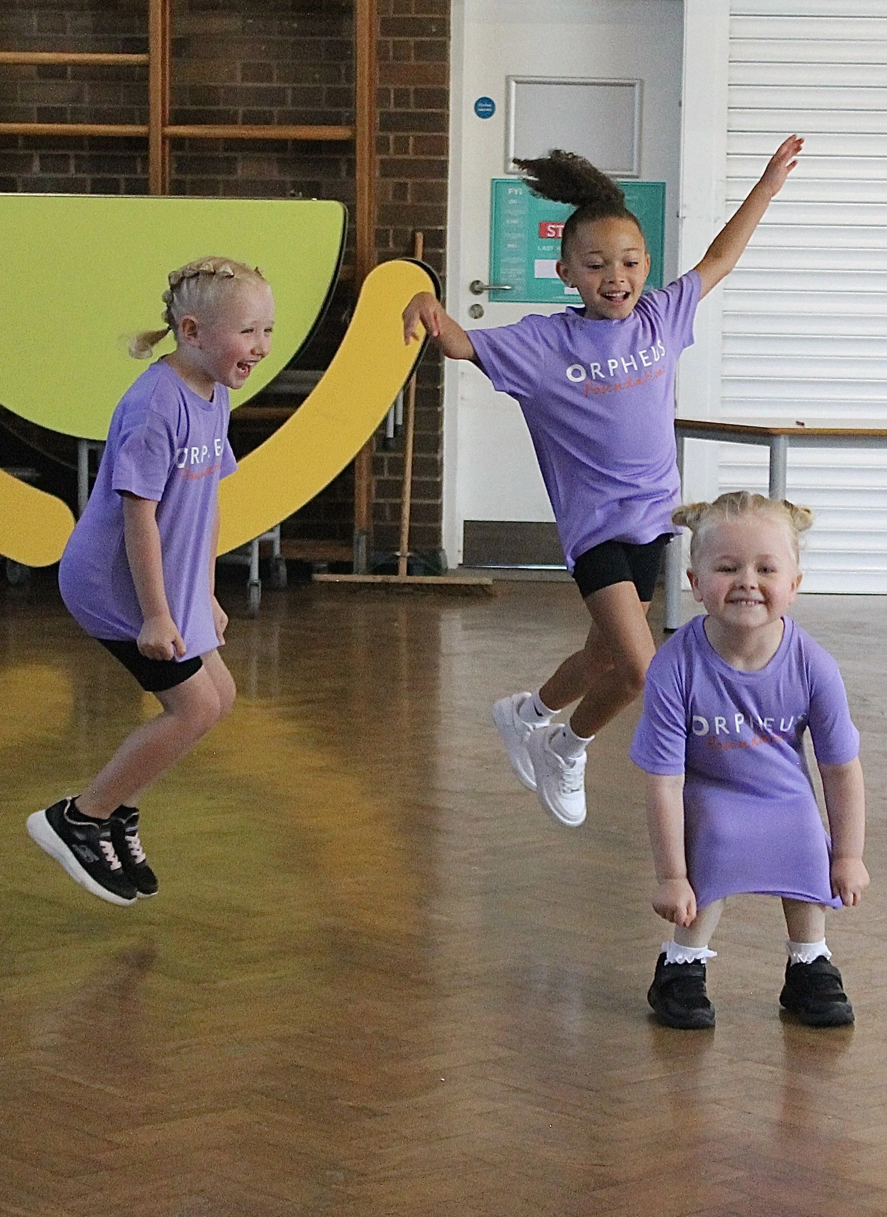 Four young girls with curly and braided hair are playing and jumping in a dance studio, wearing purple shirts with the word 'Orpheus' on them, and black shoes. The studio has a wooden floor, a yellow and green slide, and a white closed door in the background.