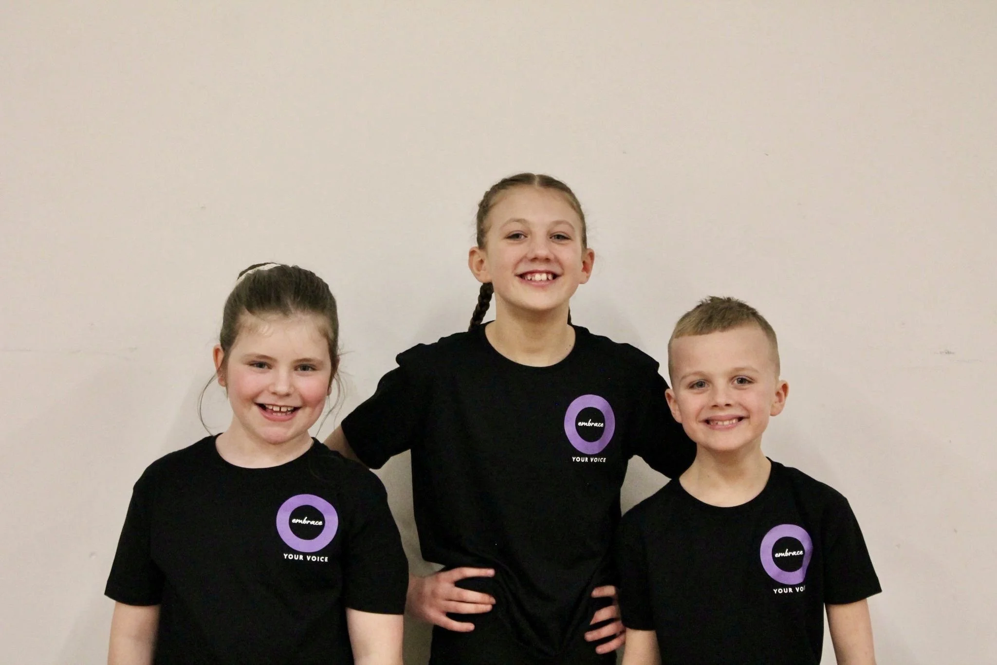 Four children standing against a plain off-white wall, wearing black t-shirts with purple circle logos and the words 'embrace your voice'.