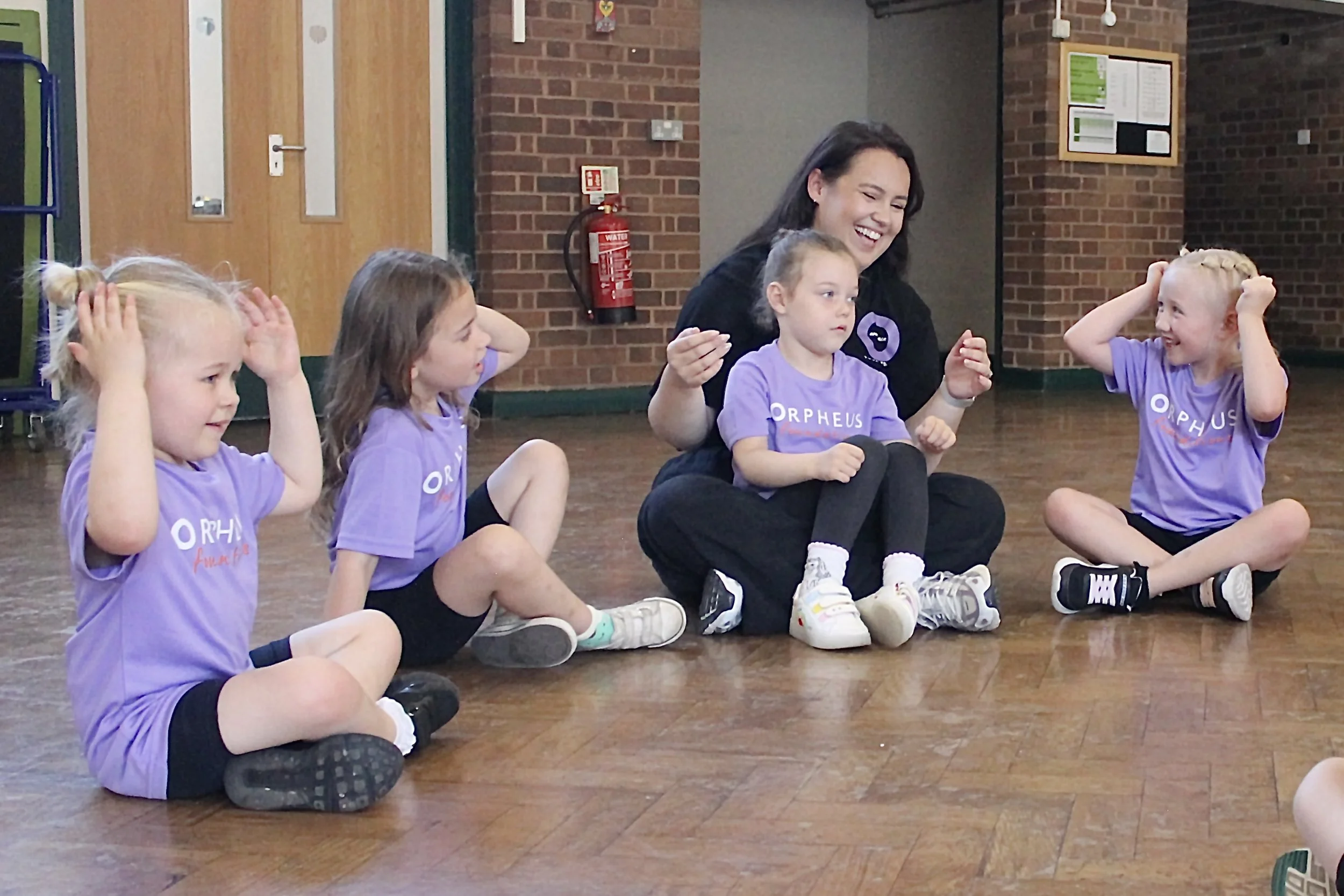 A group of young girls and their teacher sitting cross-legged on the floor in a classroom, all wearing purple T-shirts with 'ORPHEUS' printed on them, engaging in activity or conversation, with a brick wall and a fire extinguisher in the background.