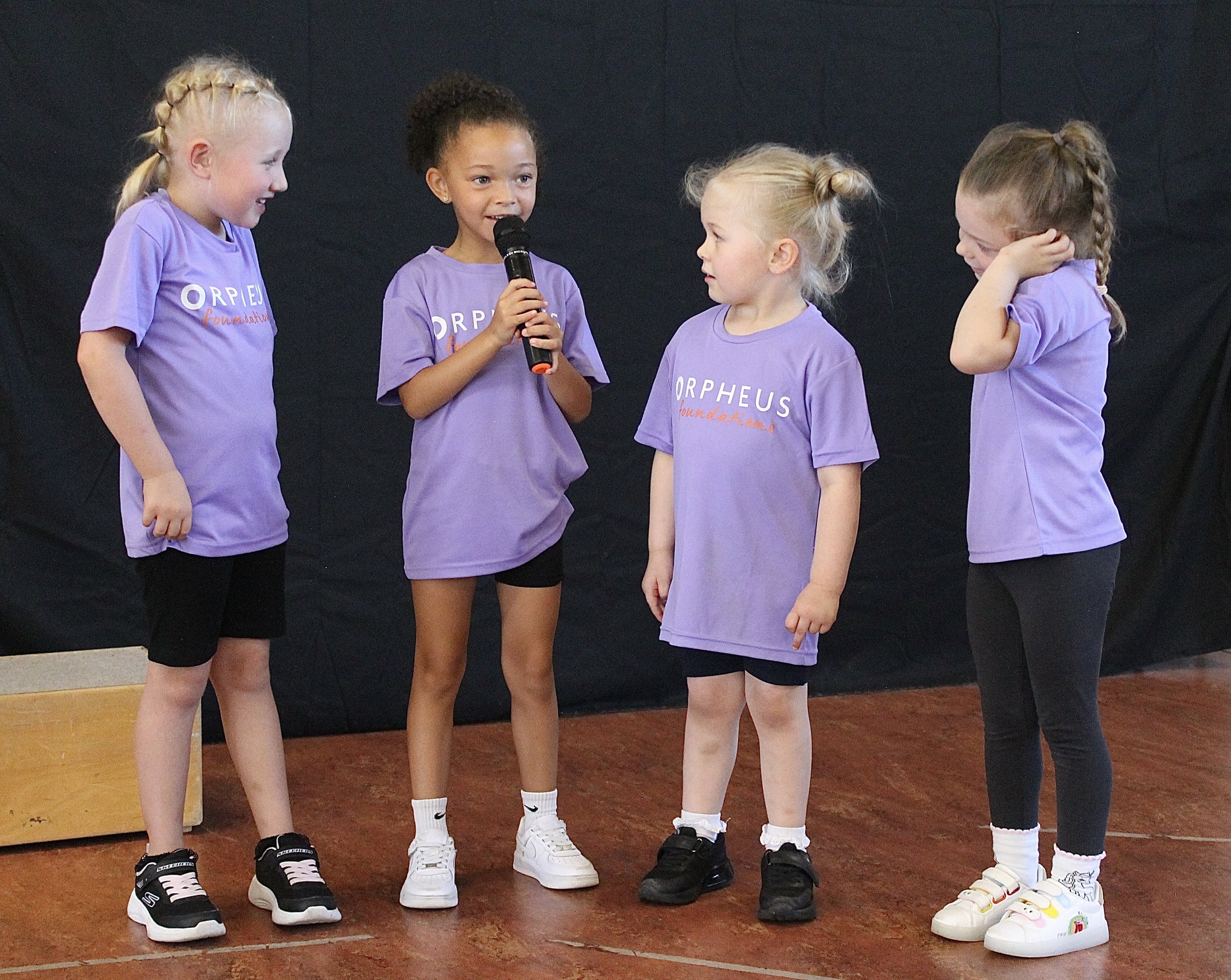 Four young girls wearing purple T-shirts with 'ORPHEUS foundation' written on them, standing on a gym floor, engaged in a conversation, with one girl holding a microphone. Their faces show smiles and attentiveness, and they are wearing sneakers and socks.
