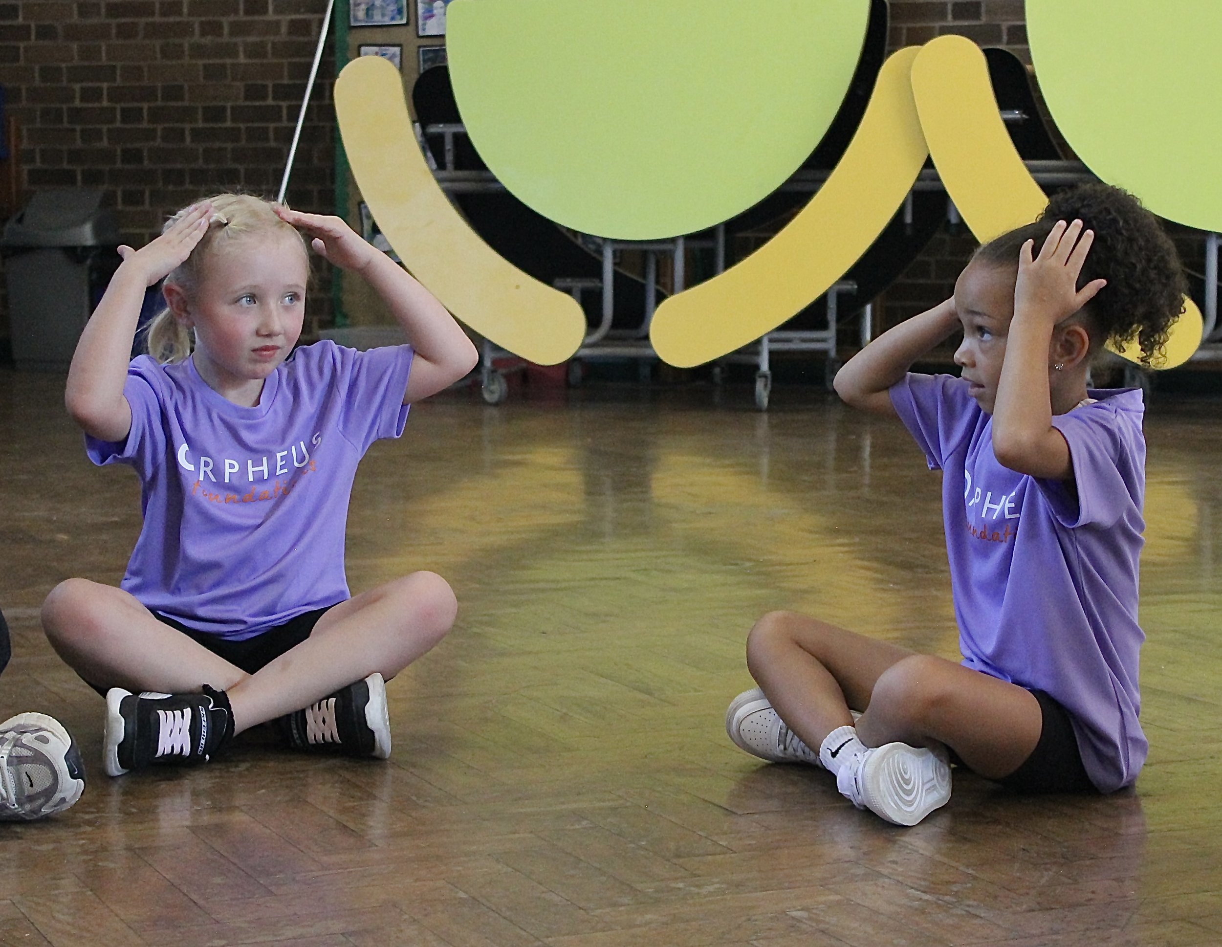 Two young girls sitting cross-legged on a wooden floor, facing each other, holding their heads with their hands, with a large colorful wall decoration behind them.