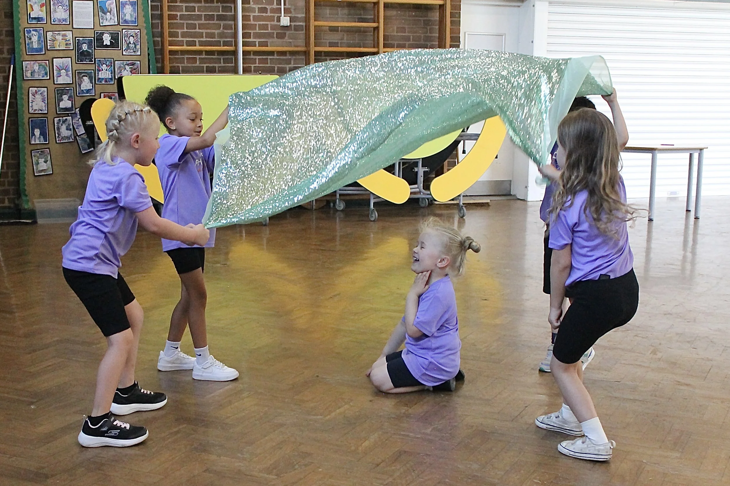 Children in a gym or classroom setting playing with a large, sparkly green fabric canopy. Some children are standing, one girl is kneeling and looking up at the canopy, and another girl is sitting on the floor, smiling with her hand on her neck.