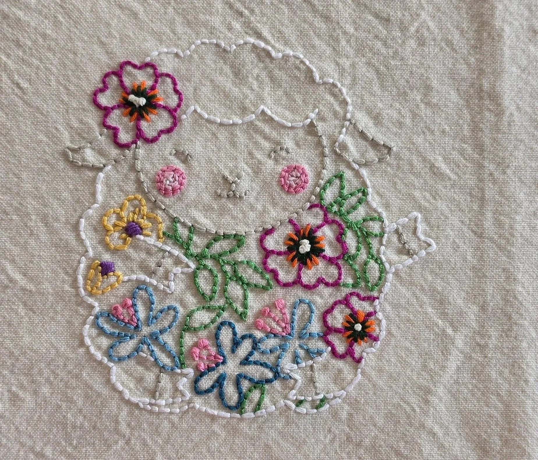 Full view of a hand-embroidered lamb surrounded by colorful flowers and green leaves on a cotton kitchen towel.