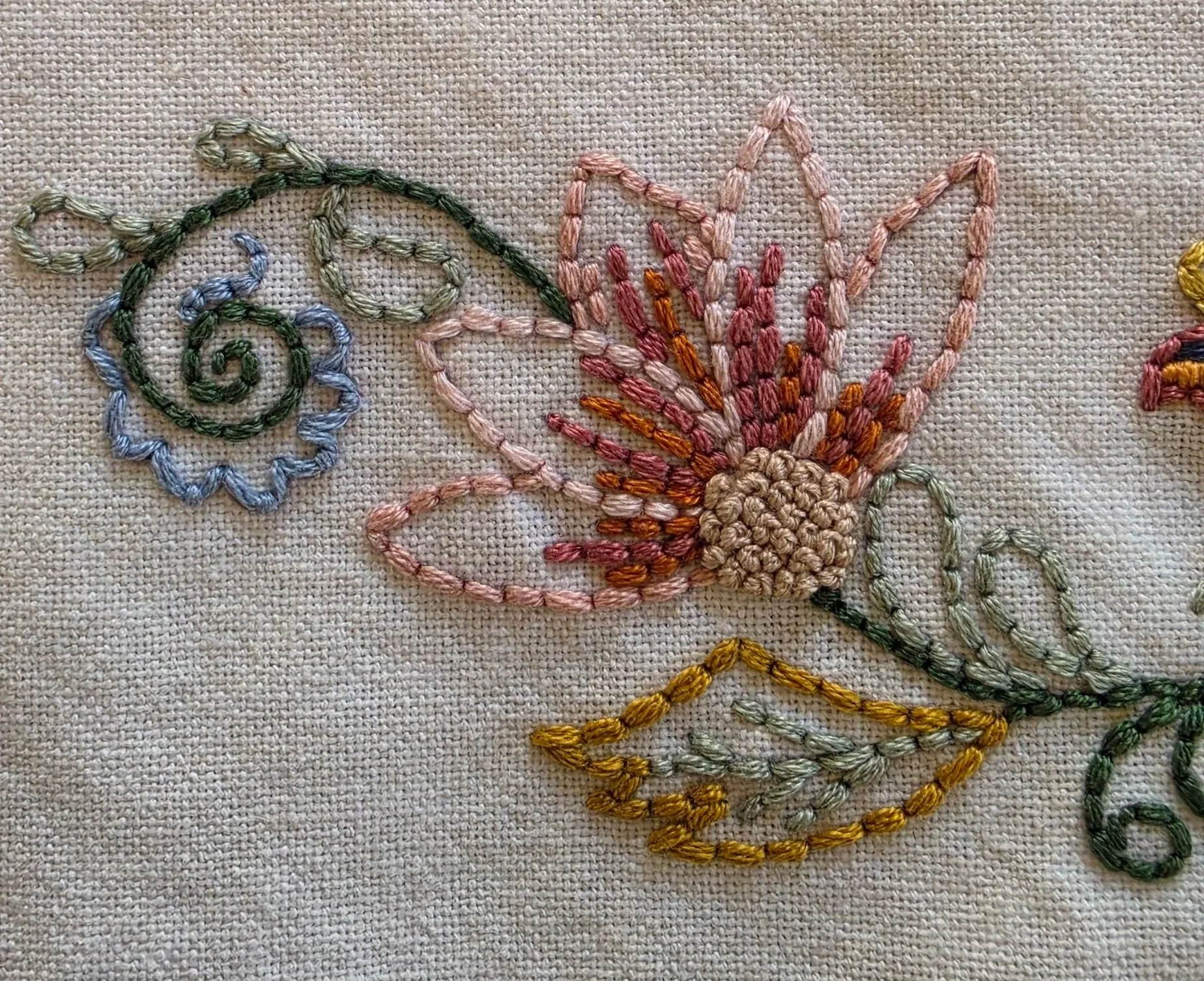 Close-up of pink embroidered flower petals and blue blossoms with green vines on cream cotton fabric.