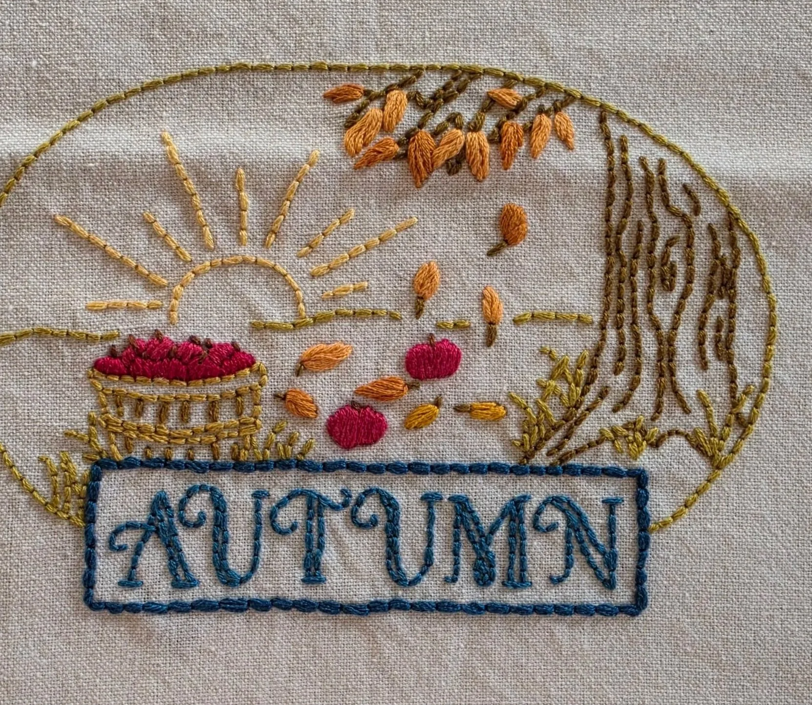 Hand-embroidered autumn harvest scene with a golden sun, red apples, falling leaves, a tree, and blue AUTUMN lettering on a beige towel.