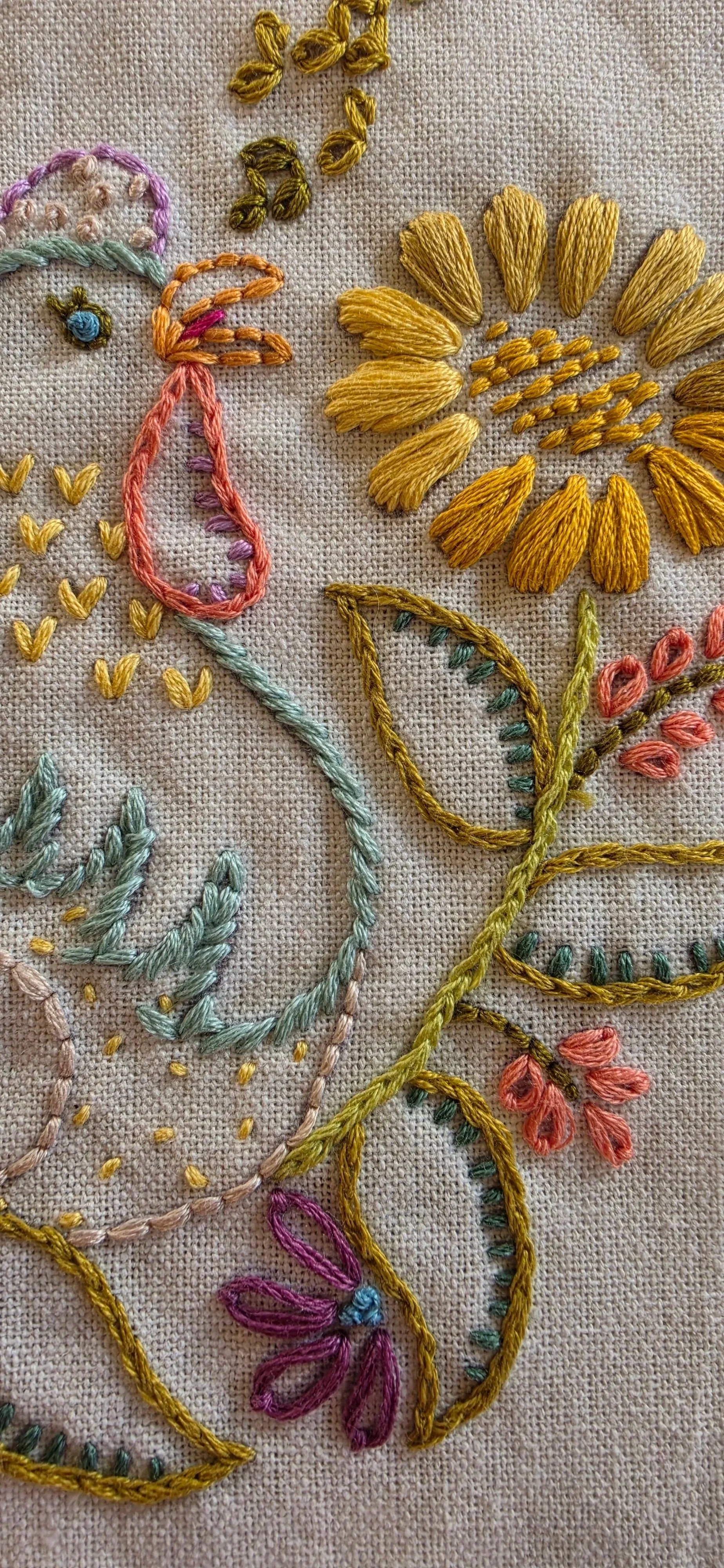 Close-up of colorful embroidered hen details, teal feathers, golden sunflower petals, and green leaves on beige fabric.