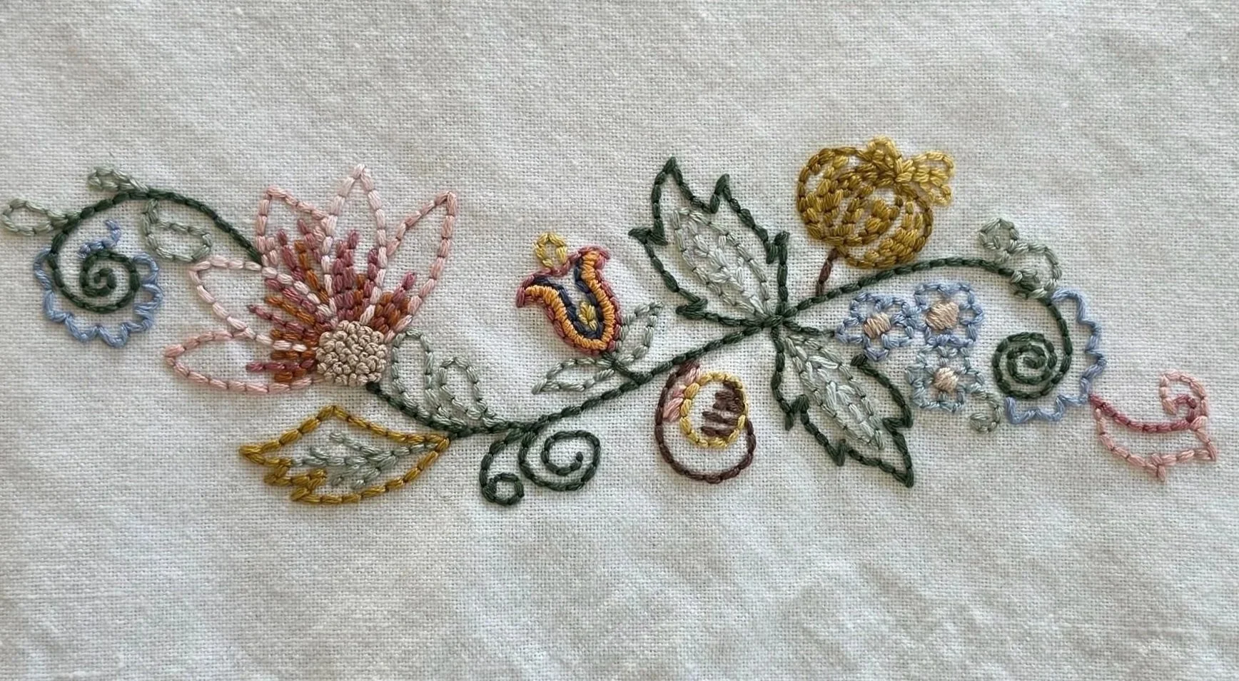 Close-up of pink embroidered flower petals and blue blossoms with green vines on cream cotton fabric.