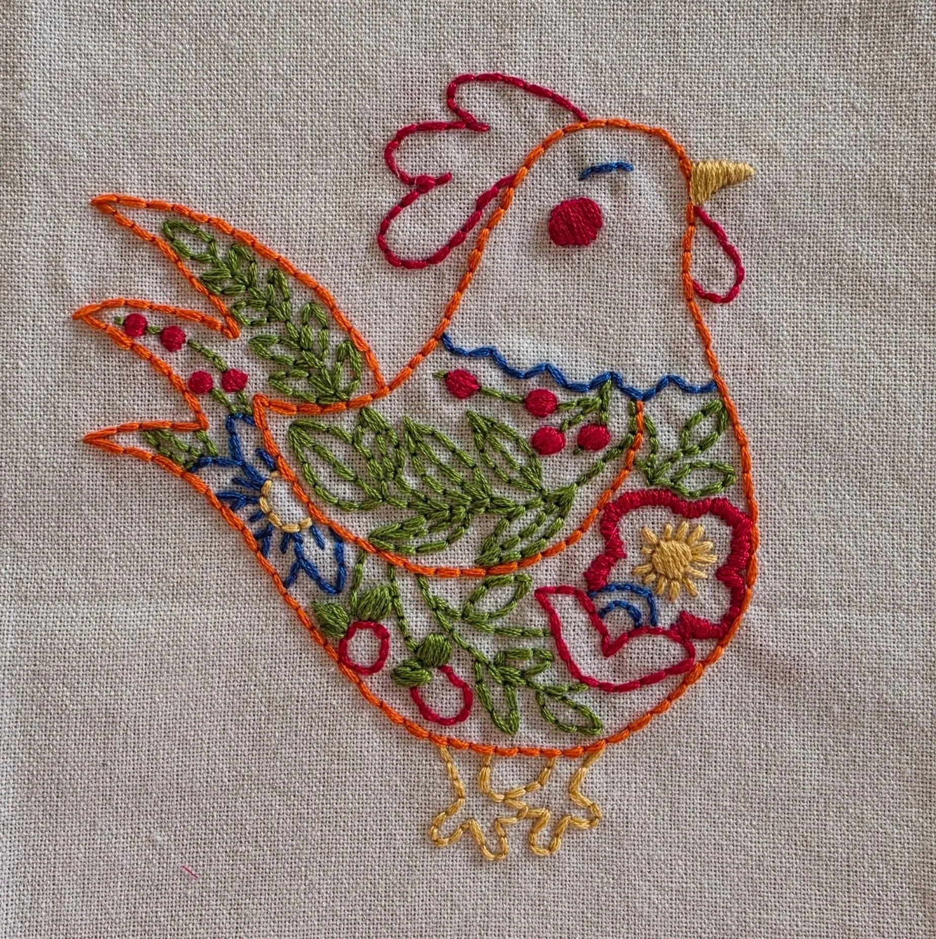 Full view of a hand-embroidered hen with a red comb and colorful flowers and patterns across its body.