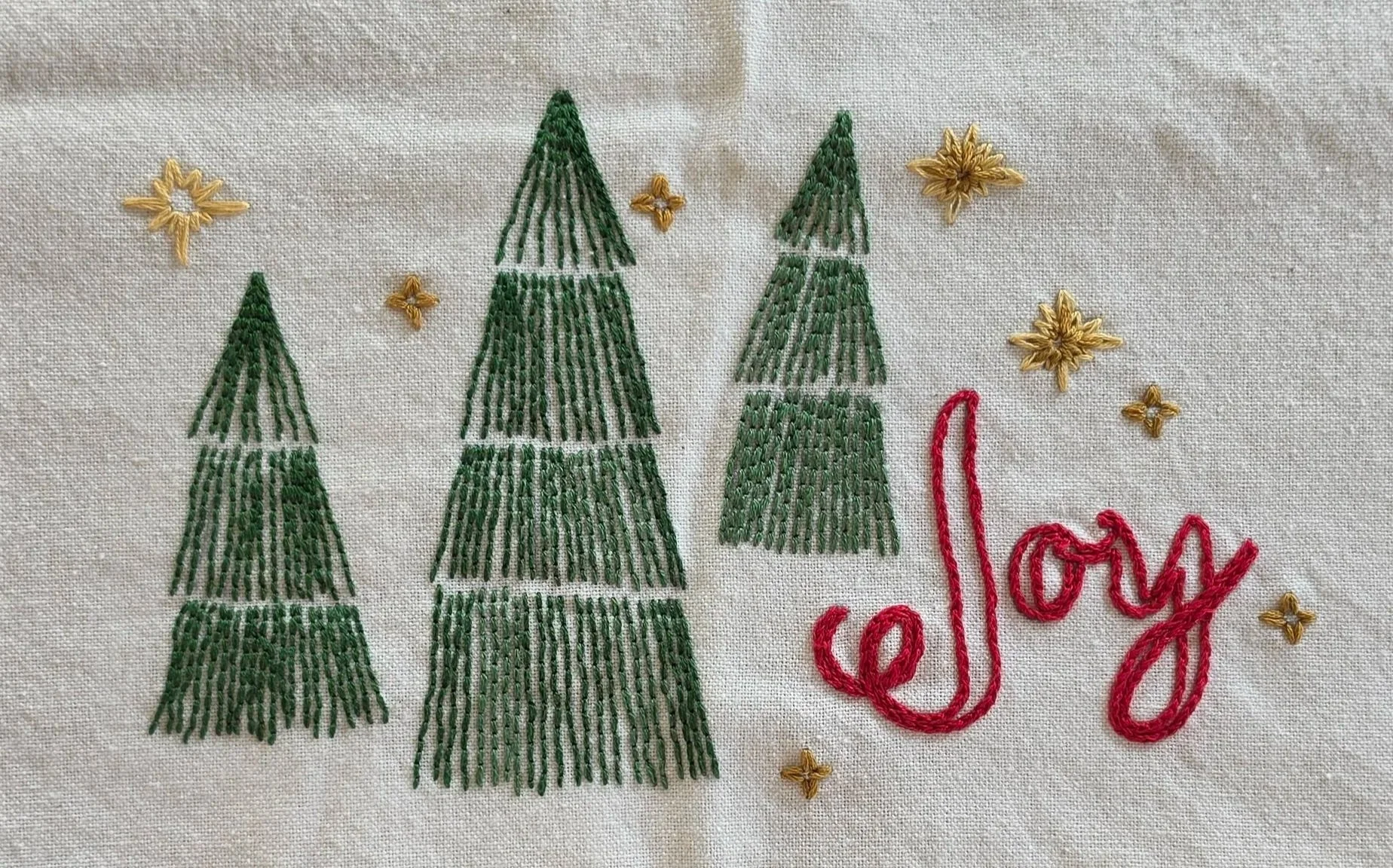 Three hand-embroidered evergreen trees beside the word Joy on a cotton Christmas kitchen towel.