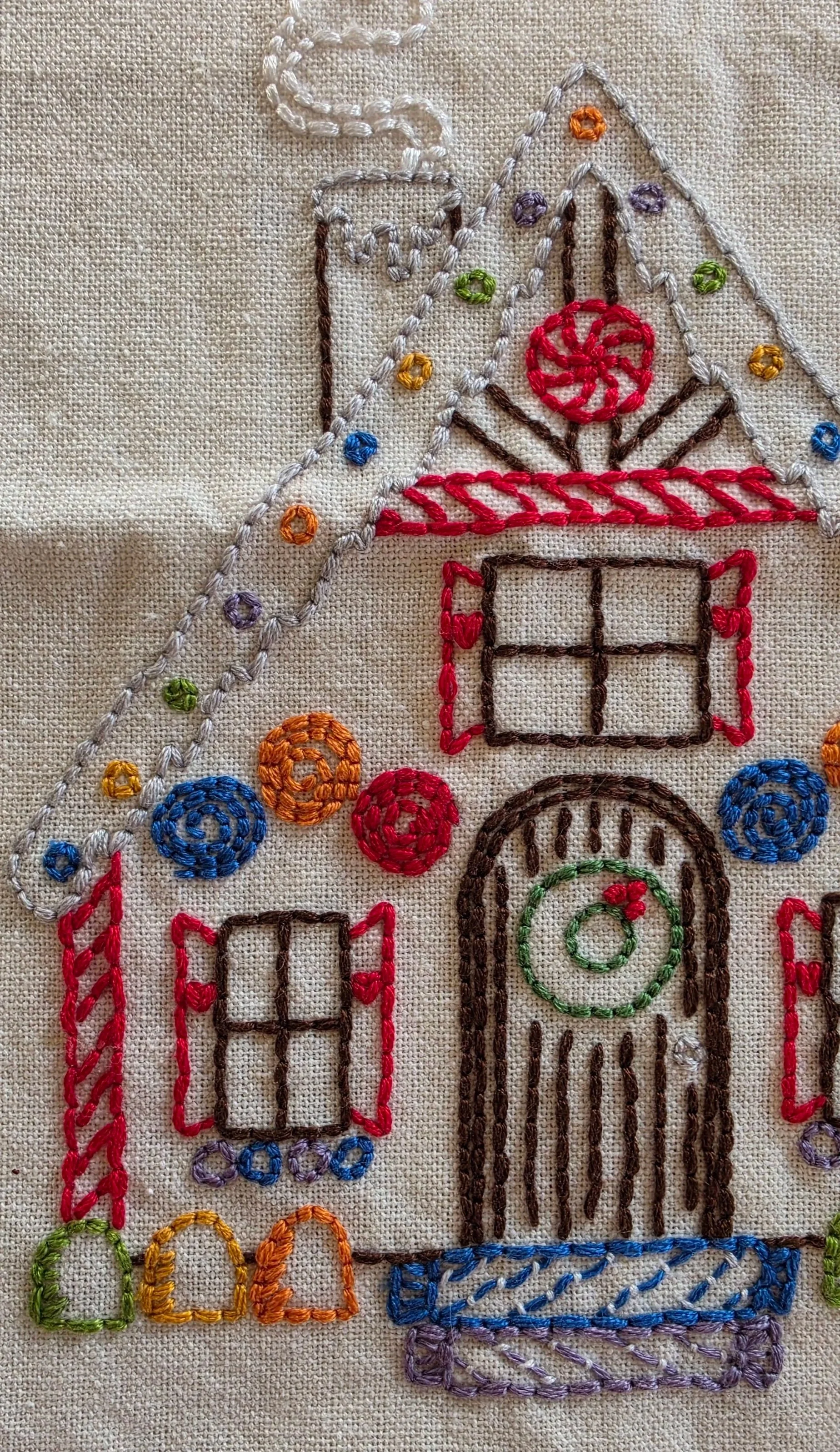 Close-up of colorful hand embroidery on a gingerbread house towel showing candy decorations, peppermint detail, windows, and a festive wreath.
