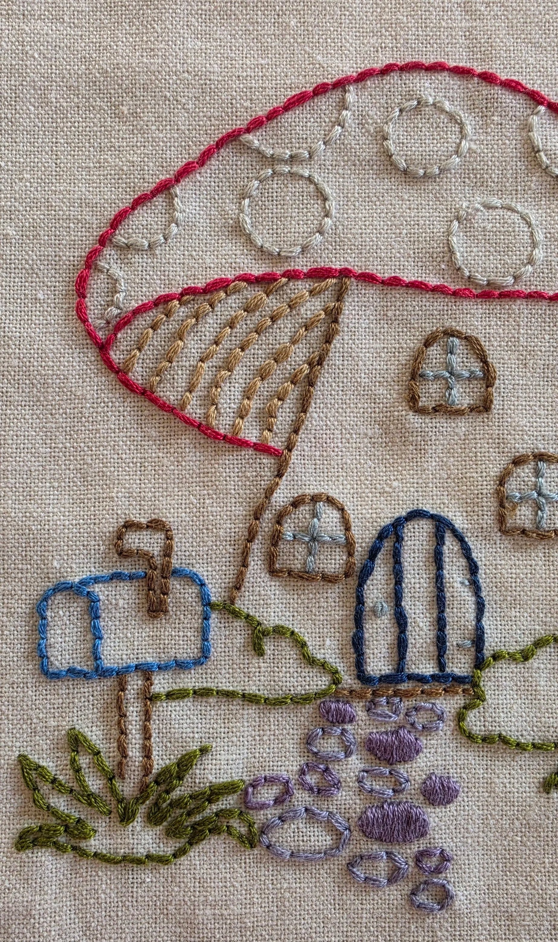 Close-up of blue embroidered garden fencing, green vines, purple flowers, and a blue chimney on a mushroom cottage towel.