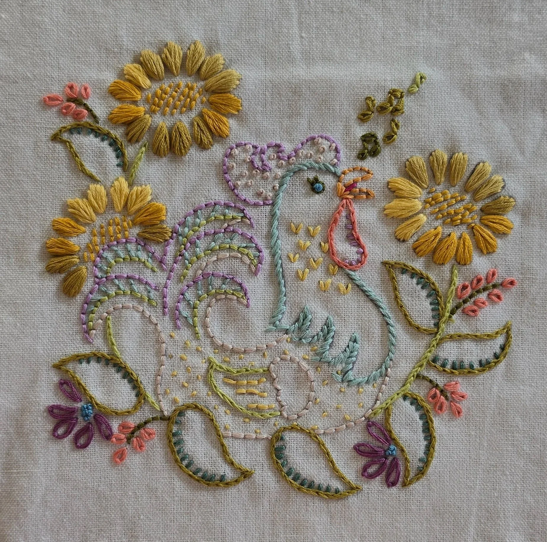 Full view of a colorful and-embroidered hen surrounded by golden sunflowers, leaves, and decorative flowers on a natural beige kitchen towel.