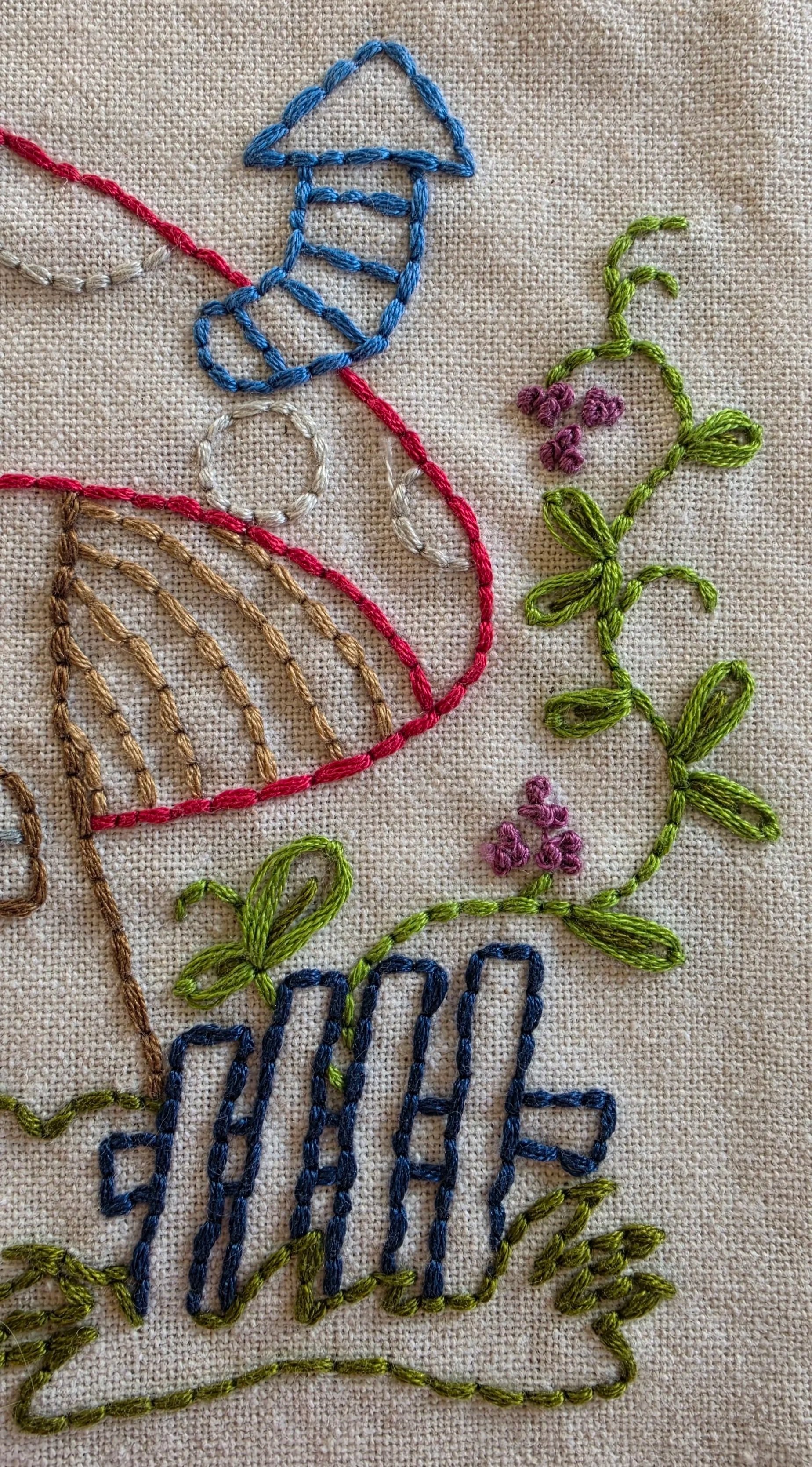 Close-up of a hand-embroidered mushroom cottage with a blue arched door, tiny windows, blue mailbox, and red mushroom roof.