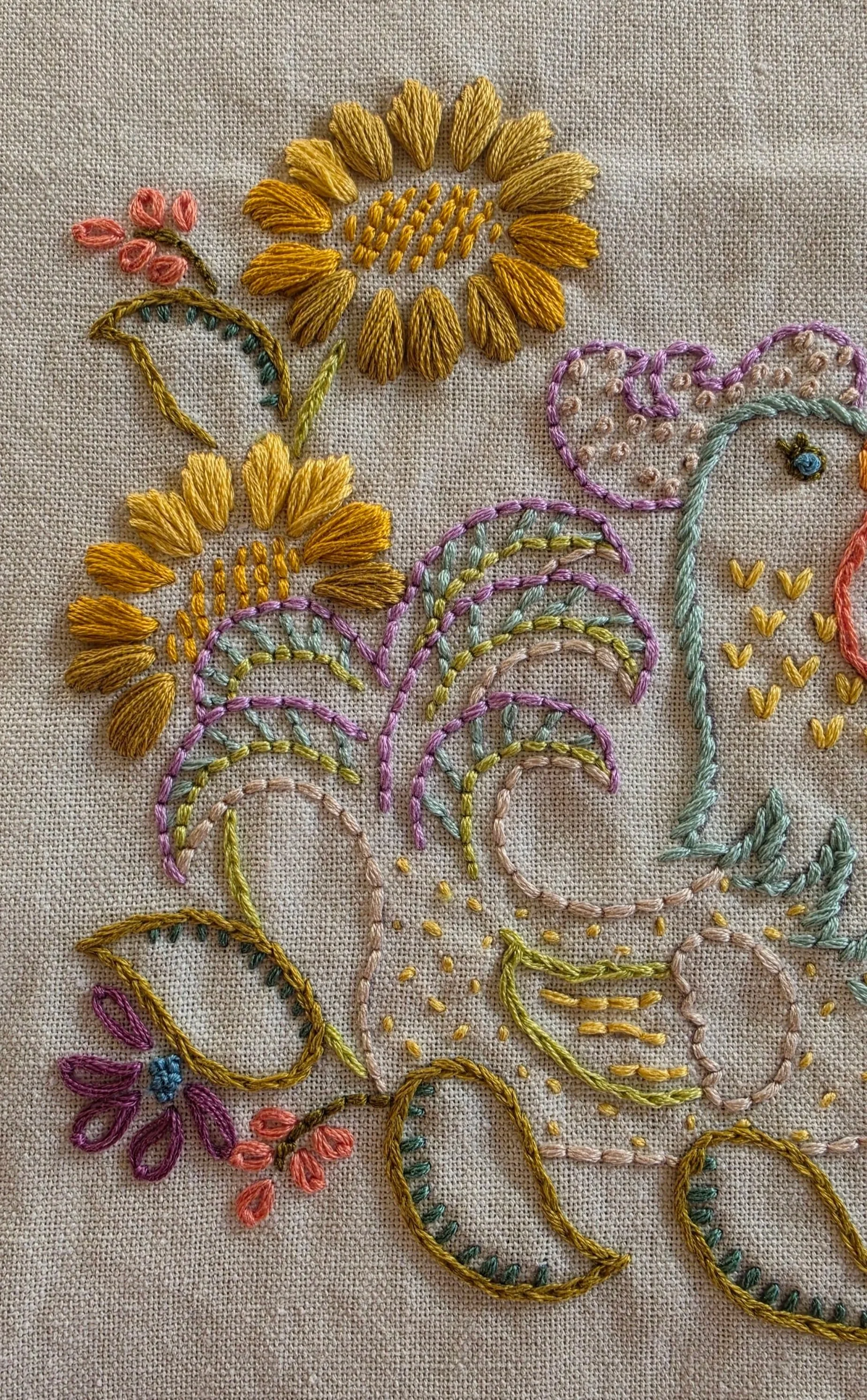 Detailed hand stitching on a farmhouse he towel, showing the hen's colorful feathers, golden sunflowers, and decorative leaves.