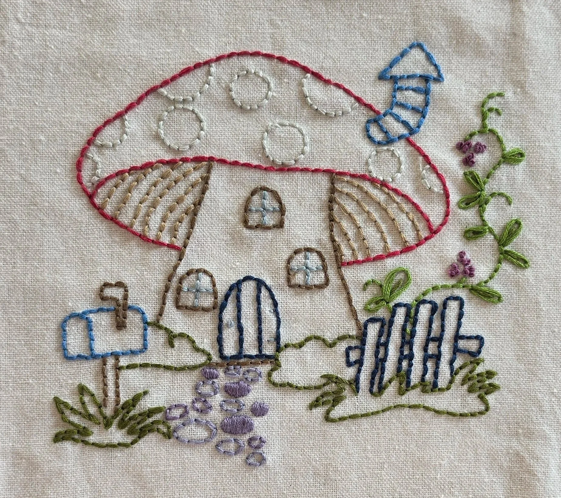 Hand-embroidered mushroom cottage with a red roof, blue door, mailbox, garden fence, and purple flowers on a natural cotton kitchen towel.