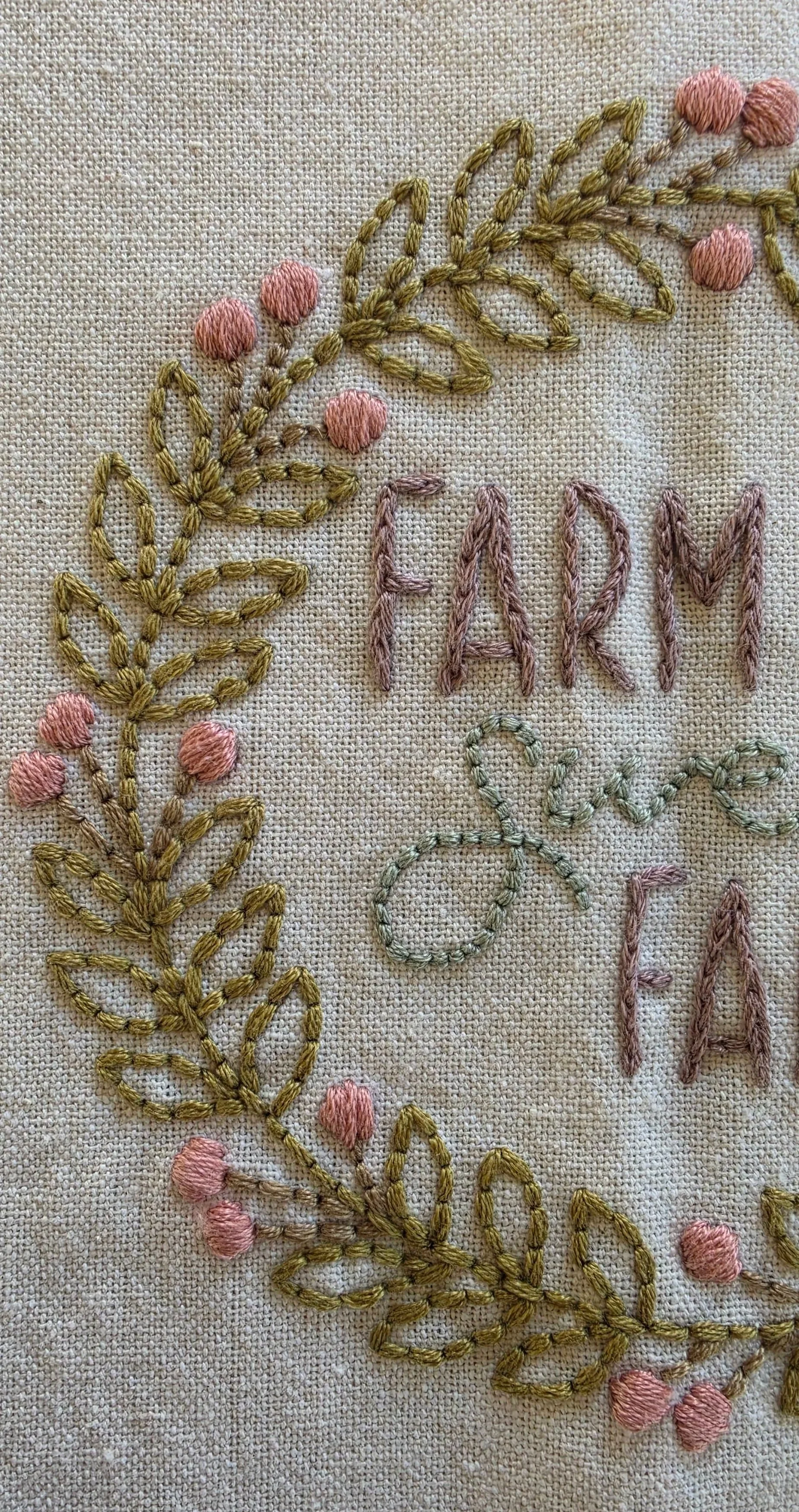 Close-up of the Farm Sweet Farm embroidered lettering surrounded by delicate pink flowers and green foliage on cream cotton fabric.