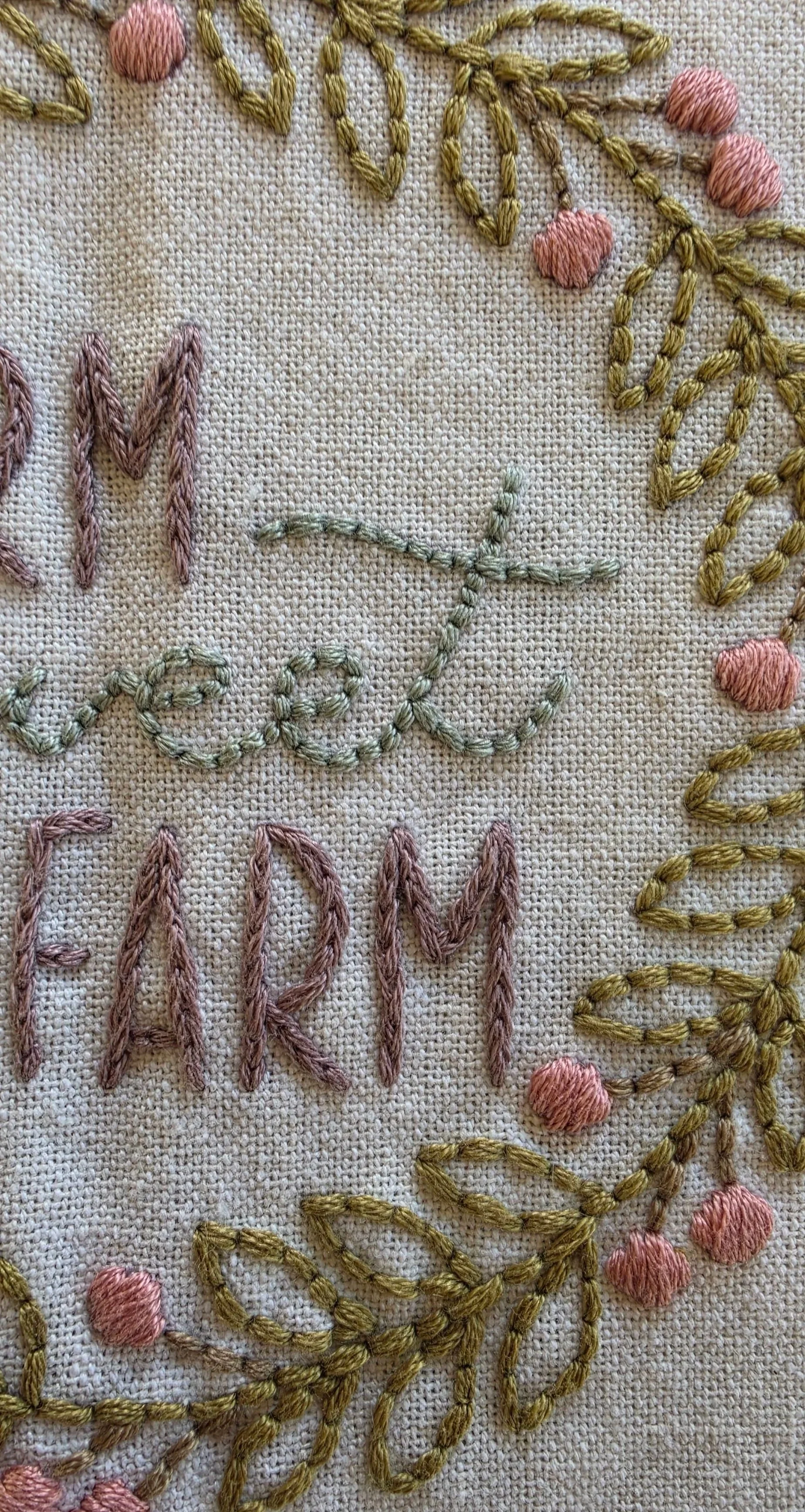 Close-up of the hand-embroidered Farm Sweet Farm lettering and decorative floral wreath, showing the colorful stitching on a cream kitchen towel.