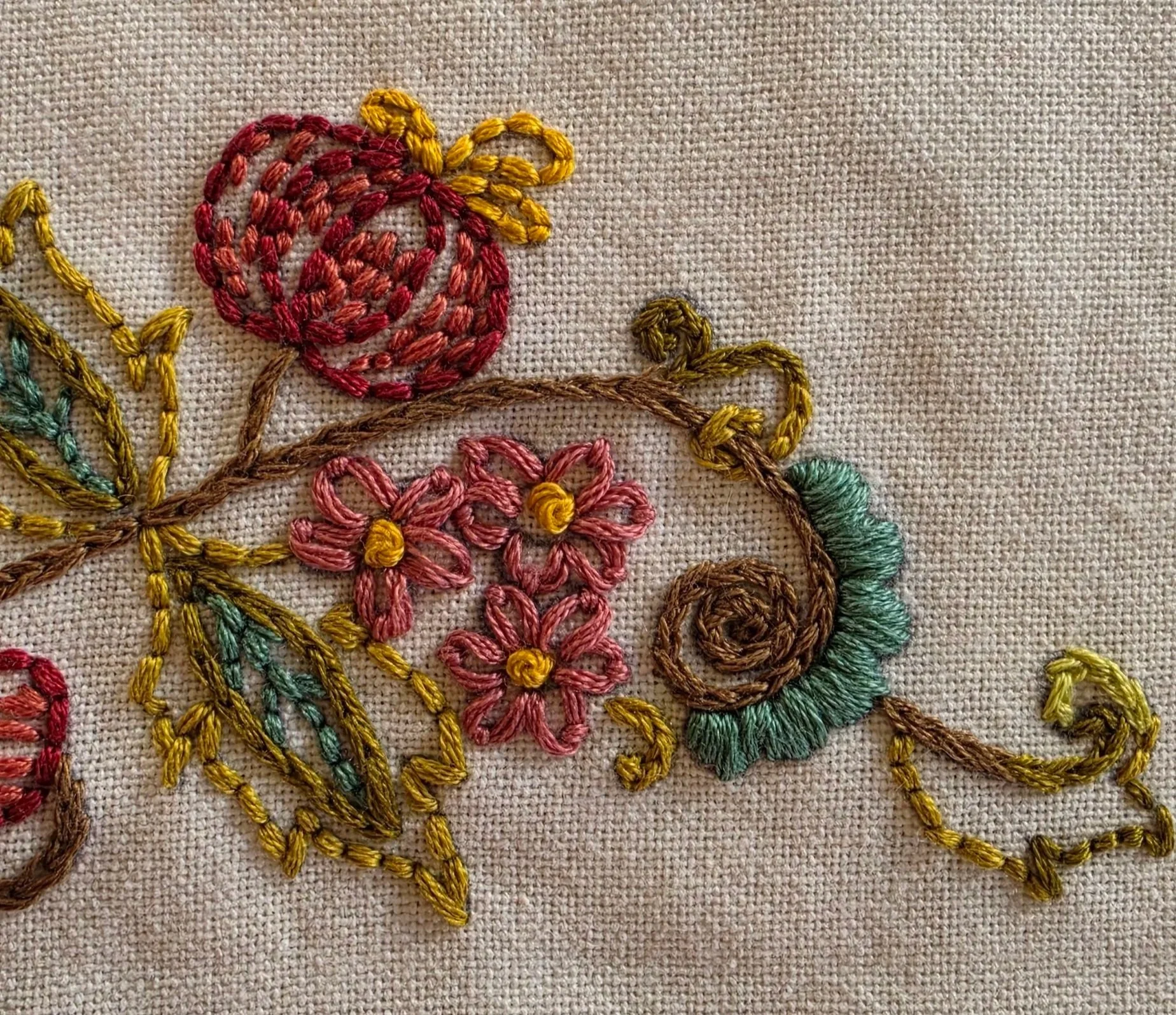 Close-up of burgundy and teal embroidered flowers with decorative green vines on cream cotton fabric.