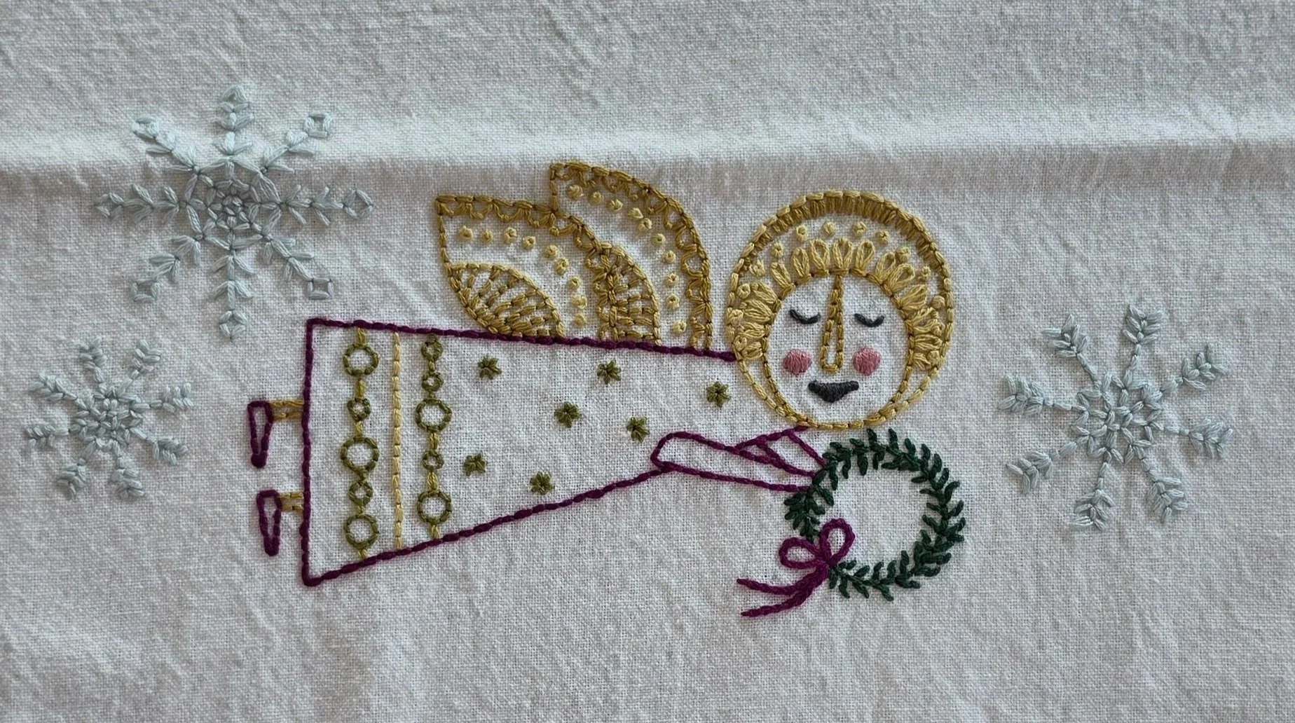 Full view of a hand-embroidered angel with a golden halo, wings, and trumpet on natural cotton fabric.