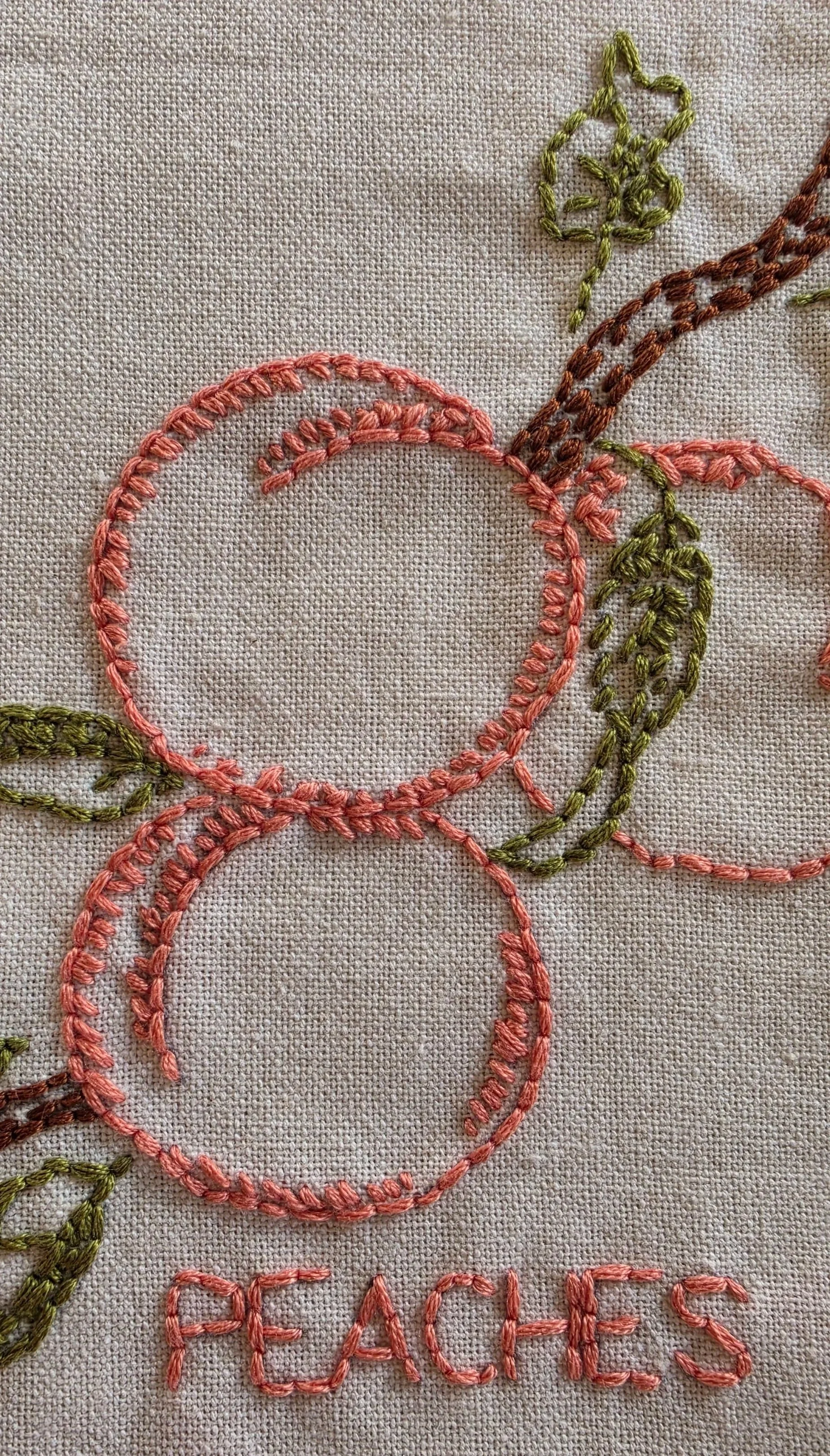 Close-up of coral peach embroidery, brown branch stitching, and green leaves on a cotton kitchen towel.