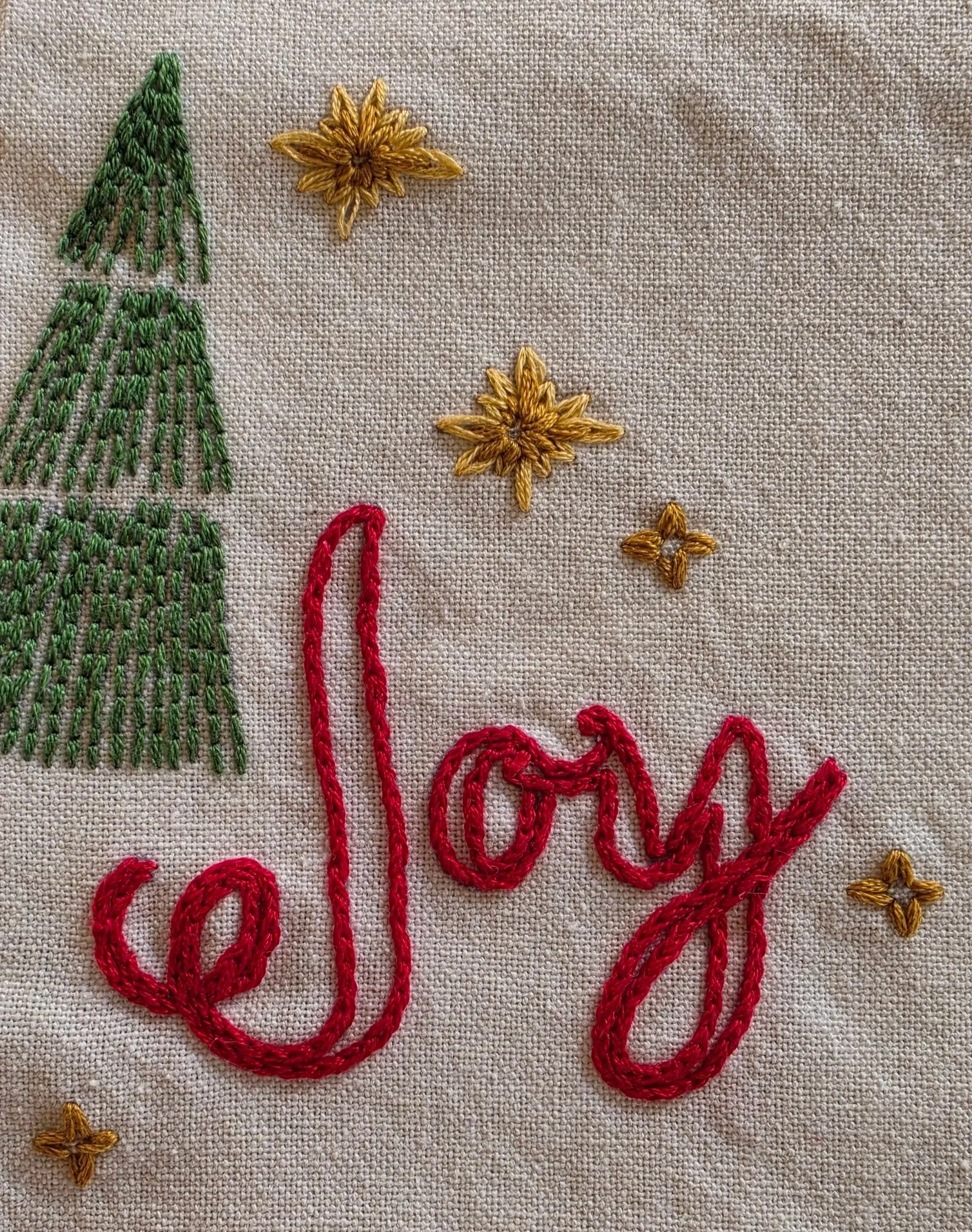 Close-up of the word Joy embroidered in decorative red script beside green evergreen trees.