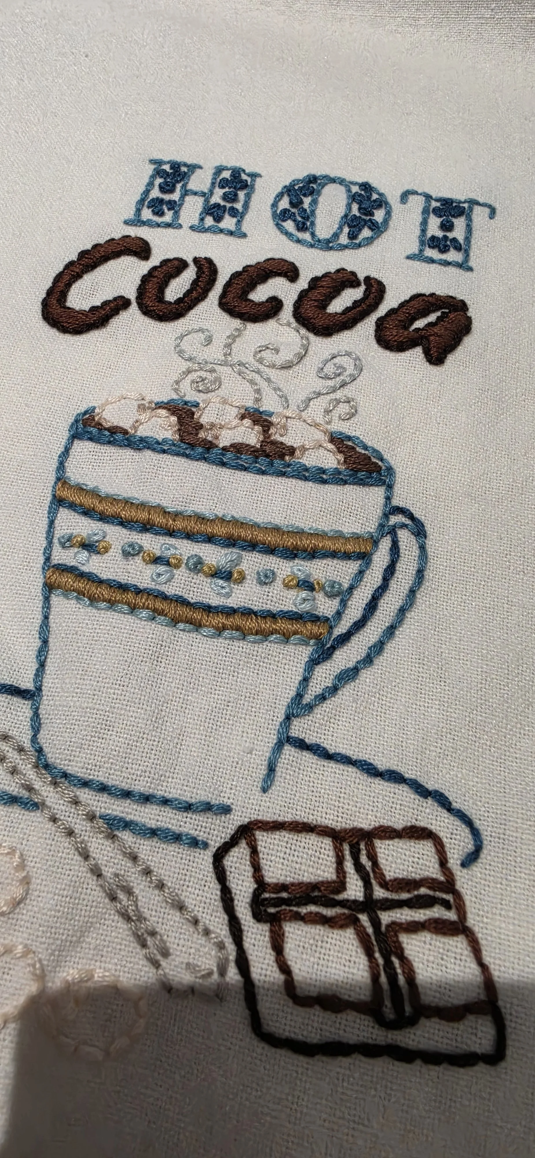 Close-up of hand embroidered hot cocoa cup with marshmallows in turquoise, gold, brown and cream thread.