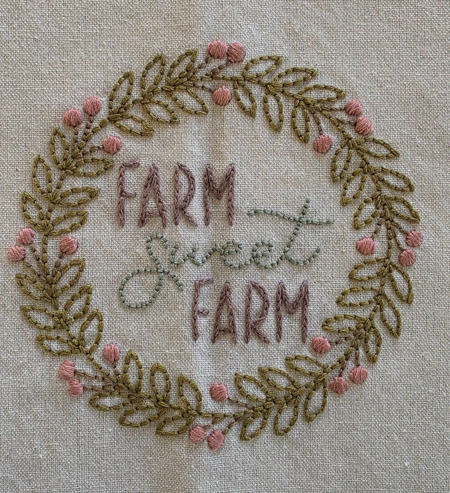 Full view of a cream cotton kitchen towel featuring the hand-embroidered words Farm Sweet Farm inside a decorative wreath of flowers and greenery.