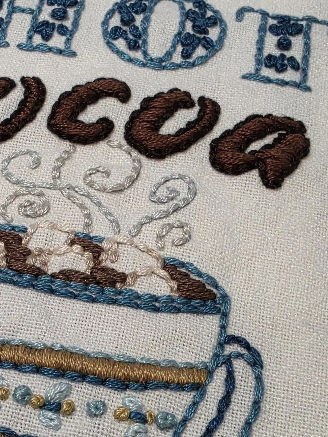 Close-up of the hand embroidered Hot Cocoa lettering and steaming mug design on a cream cotton kitchen towel.