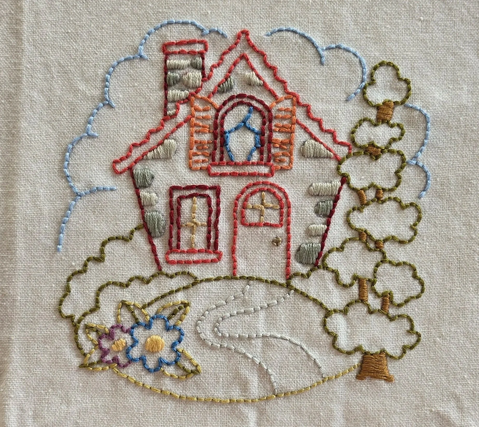 Full view of a hand-embroidered storybook cottage with a red roof, blue windows, green trees, winding path, and flowers.