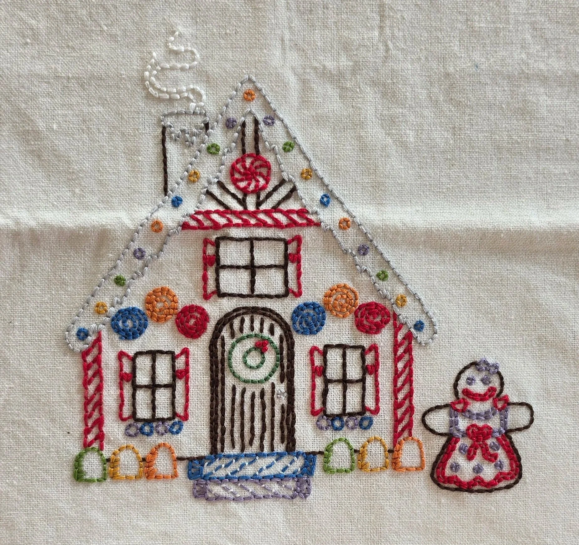 Full view of a hand-embroidered gingerbread house with colorful candy decorations, a wreath on the door, and a gingerbread girl on a natural cotton towel.