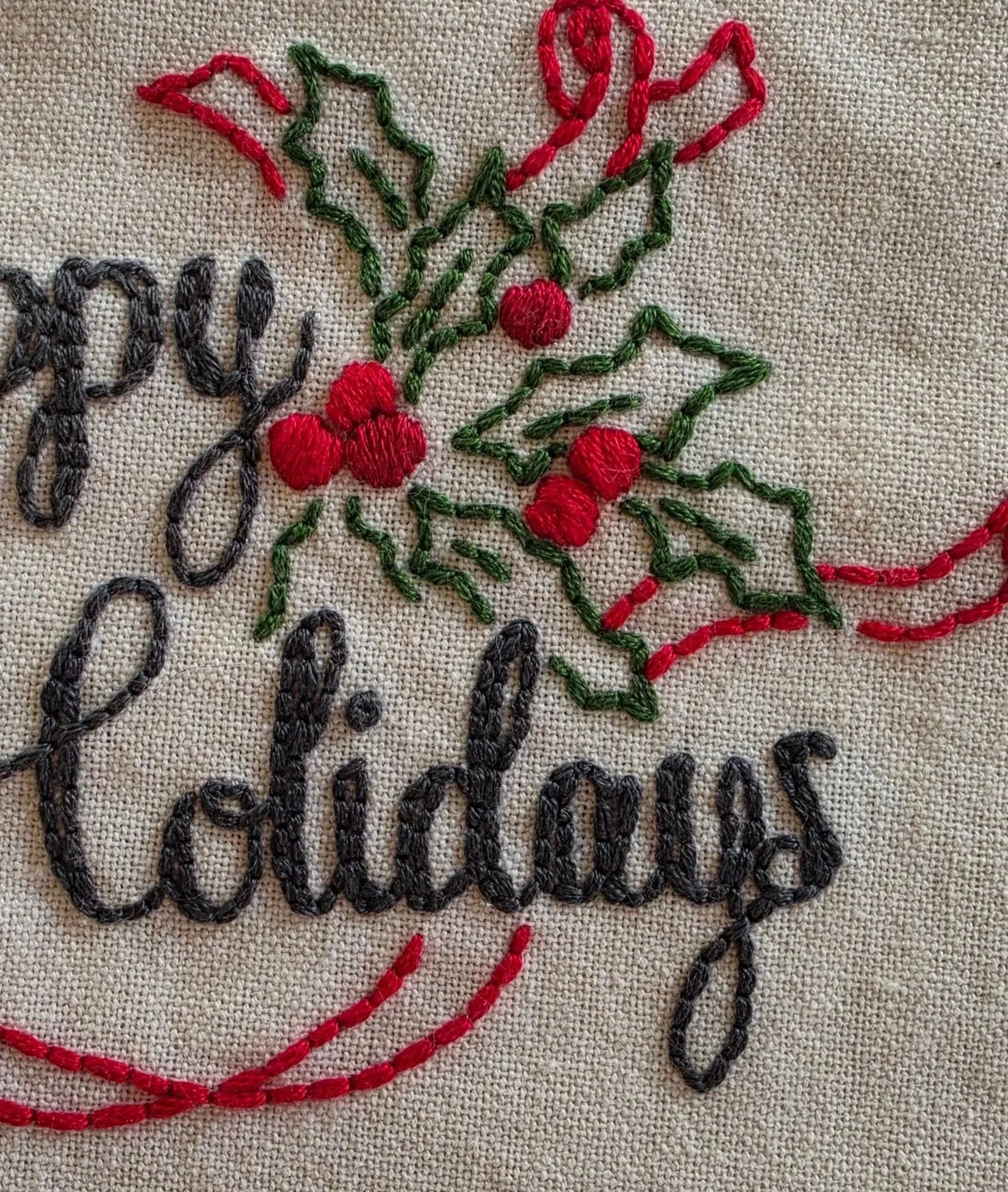 Close-up of Happy Holidays script embroidery with holly leaves and red berries stitched on cotton fabric.